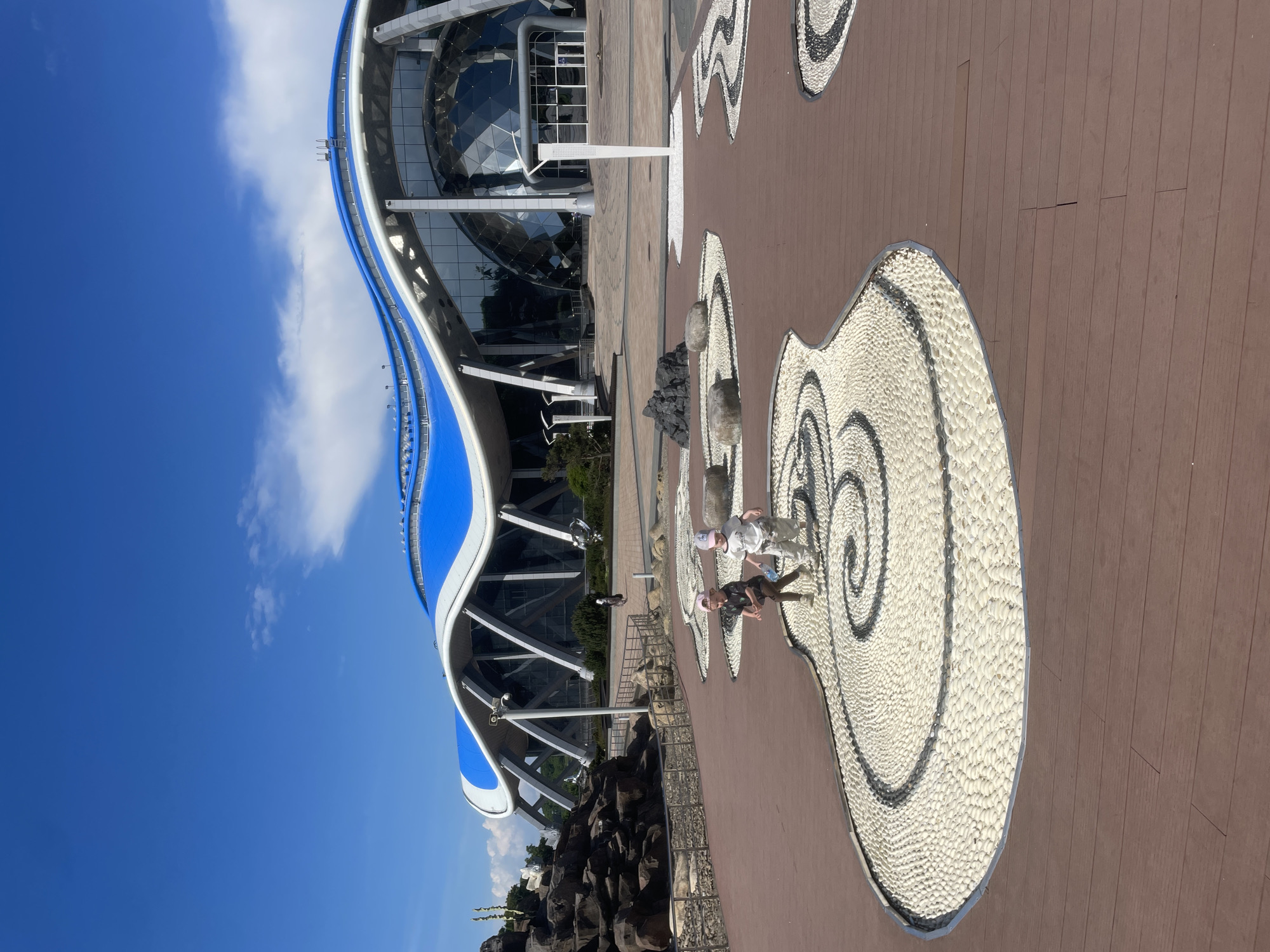 In the city of Russky, Russia, two young children are captured in mid-stride on a circular mosaic pathway. The pathway, made of white pebbles and dark stone accents forming a spiral pattern, is bordered by dark wooden planks. In the background, a modern building with a distinctive undulating blue and white roof and large glass panels stands under a bright blue sky with scattered clouds. The scene also features a rocky landscape on the left and a few large boulders placed on the pathway.
