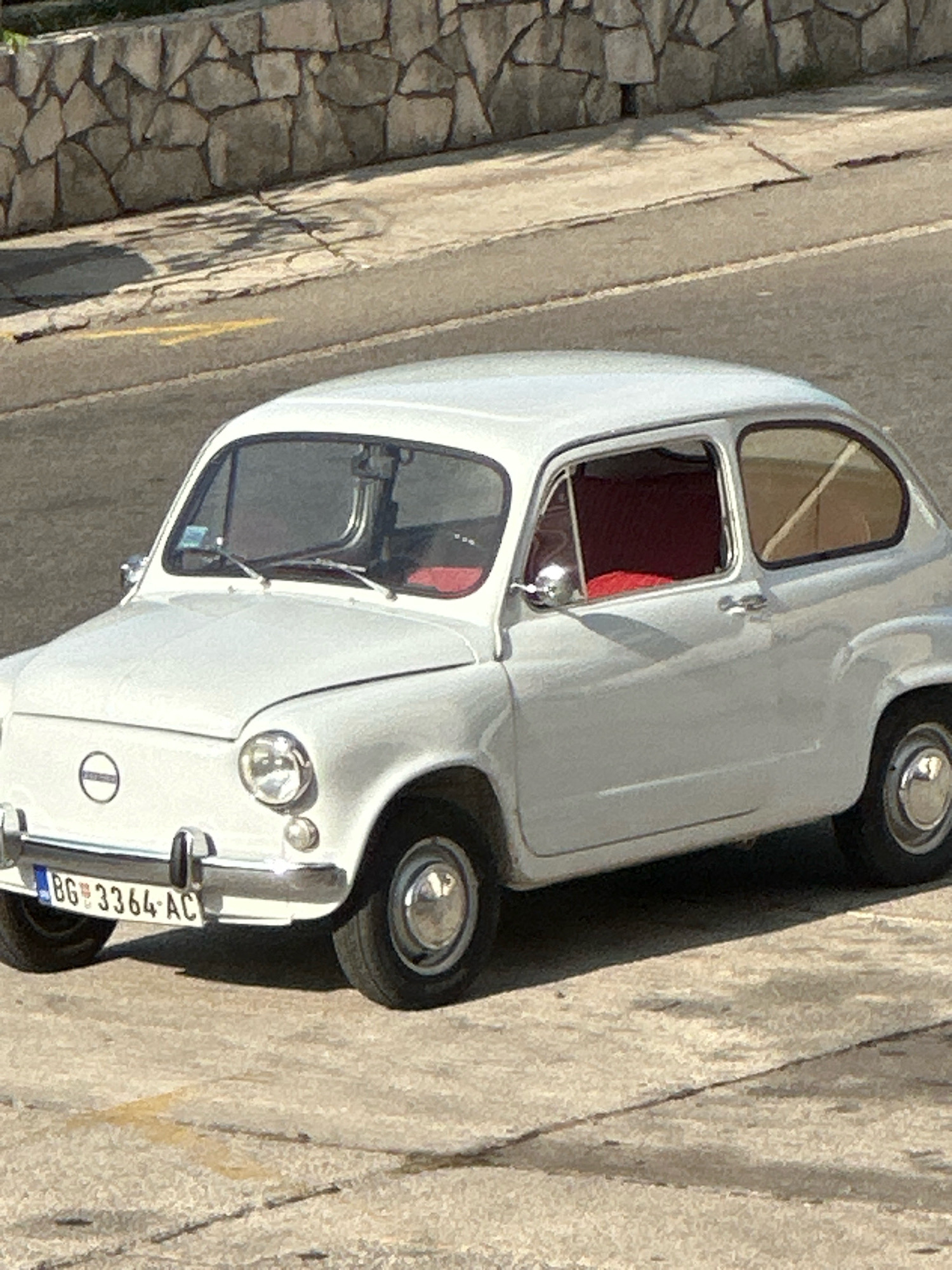 An abandoned white Fiat 600 is parked on a cracked concrete surface. The car has red interior and a license plate that reads "BG 3364-AC". Behind the car, a stone wall and a street are visible, suggesting a setting in the city of Budva, Montenegro. The image is captured in daylight, with shadows suggesting a sunny day.
