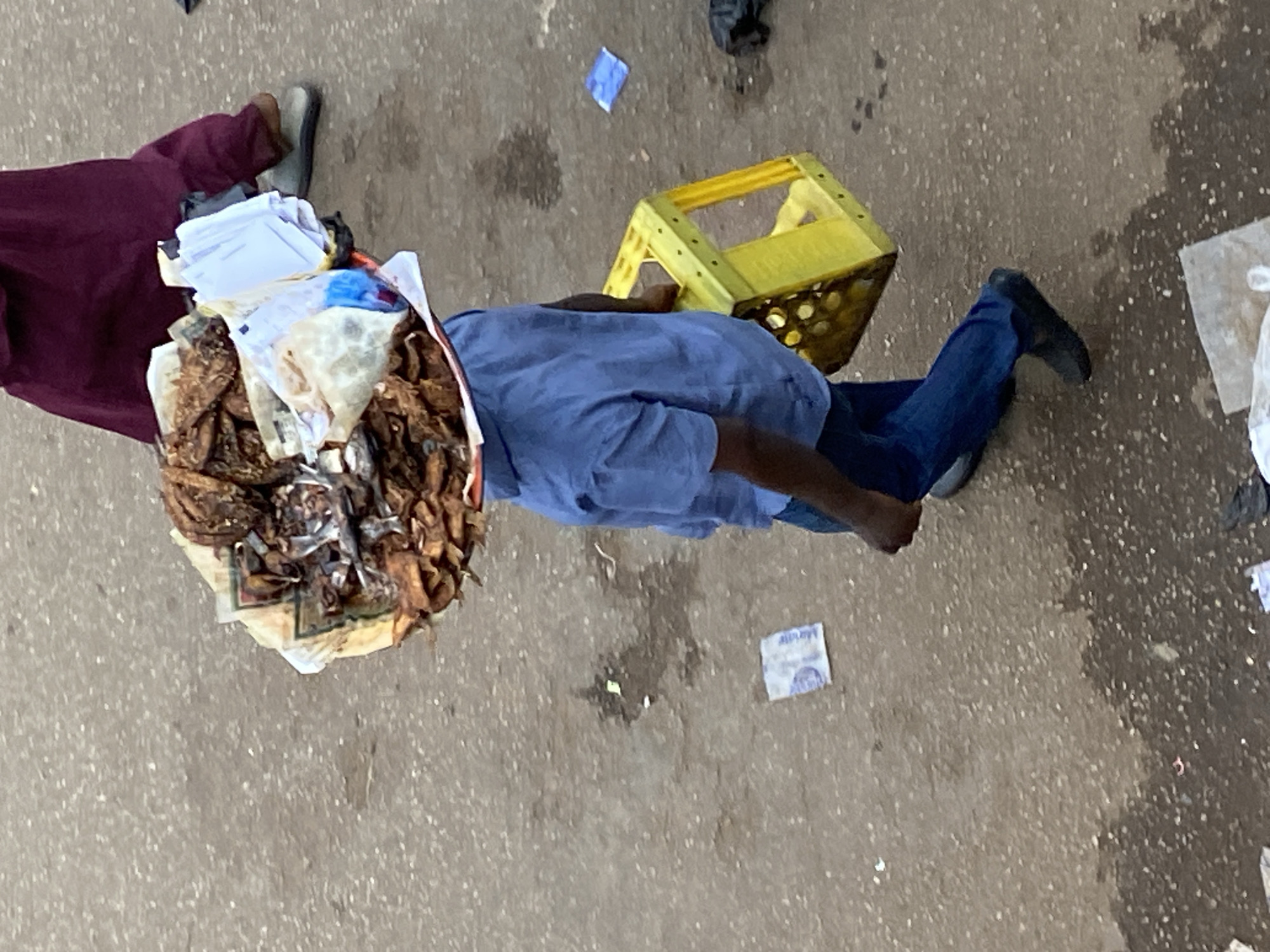 This overhead image captures an outdoor scene in Bukuru, Nigeria, likely during the daytime on a paved surface such as a street or market. The ground is stained and scattered with small pieces of litter.
The central subject is a person lying flat on their back on the ground, wearing a light blue short-sleeved shirt, blue jeans, and black shoes. Their face is not visible. Next to their head, towards the upper left, is a large, irregularly shaped mound of what appears to be dried fish or a similar dried food product, wrapped in various papers and plastics, suggesting it is merchandise.
Partially visible on the far left of the frame is another individual's lower body, dressed in maroon trousers and a single grey shoe. A bright yellow plastic crate is situated near the chest of the person lying on the ground.
Several pieces of debris are scattered on the pavement, including a blue wrapper and a white wrapper with some partially legible text, possibly "MARAF," alongside a faint logo. The scene depicts a moment in a public space, possibly involving a street vendor and their goods, with one individual resting or in an uncertain state on the ground.