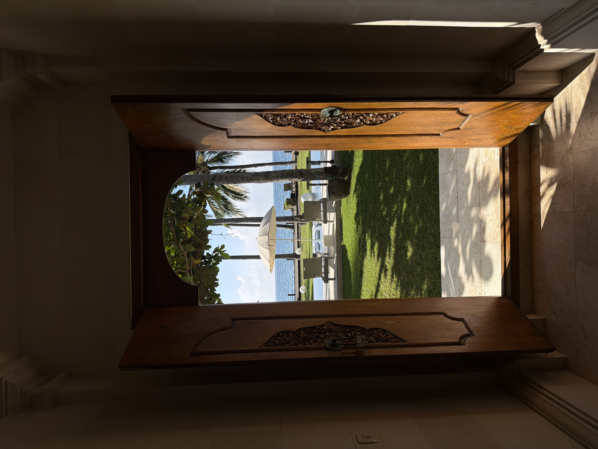 An eye-level shot taken from within a building looks out through a partially open, ornate wooden door. The door appears to lead to an outdoor area, likely a garden or patio, with a view of the ocean. The door is detailed with decorative carvings. The scene beyond the door includes a grassy area, palm trees, beach umbrellas, lounge chairs, and the ocean. The lighting suggests it is daytime, with shadows cast by the trees and building structure. The location is Karangasem, Indonesia.