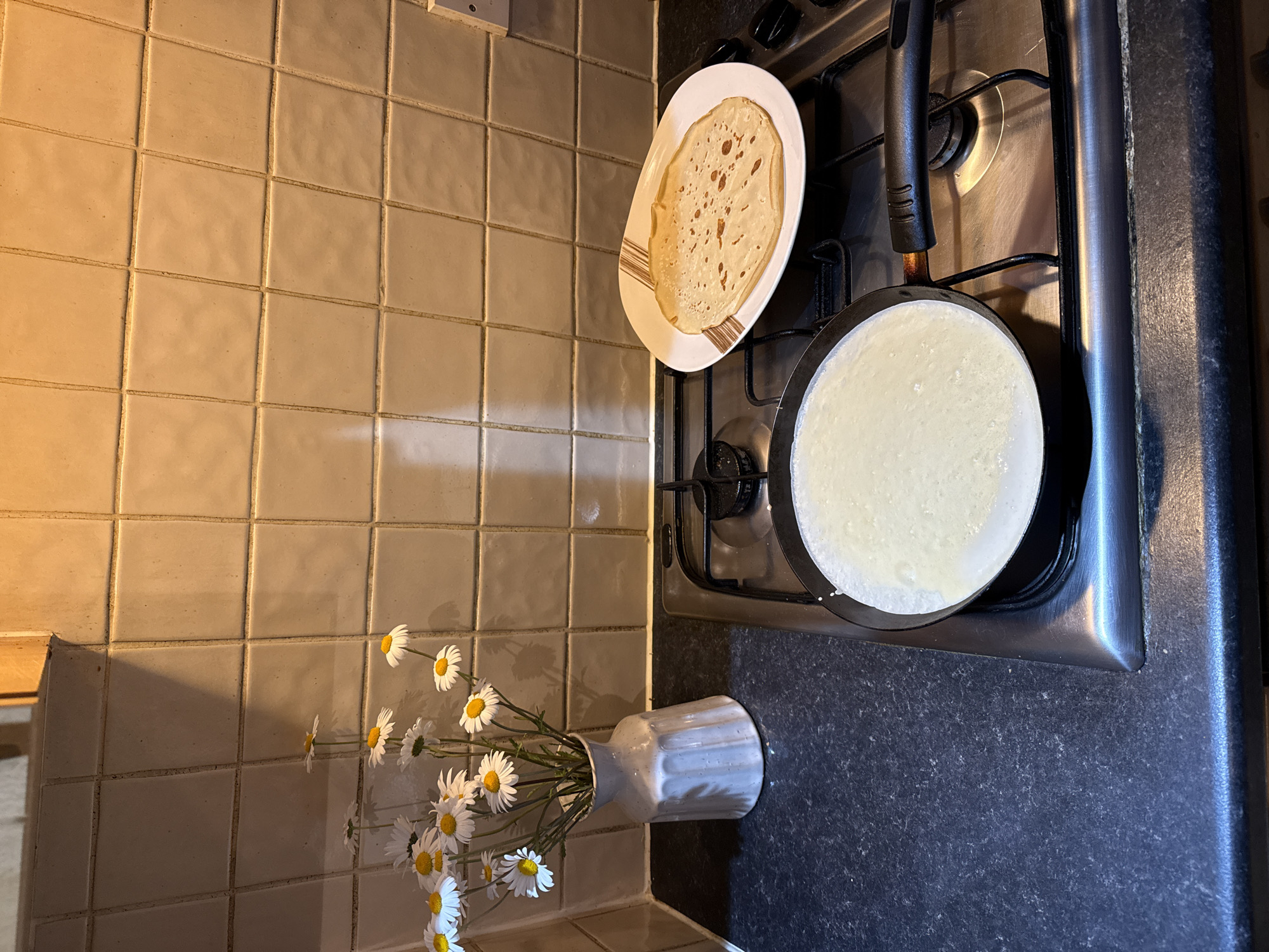 A kitchen scene in Great Yarmouth, United Kingdom, features a gas stove with a black pan containing a pale, uncooked pancake batter. Next to the pan, on the stove, lies a plate with a cooked pancake. Adjacent to the stove, a white, textured vase holds a bouquet of white daisies. The backdrop is a wall covered in light-colored, square tiles. The countertop is dark and textured.