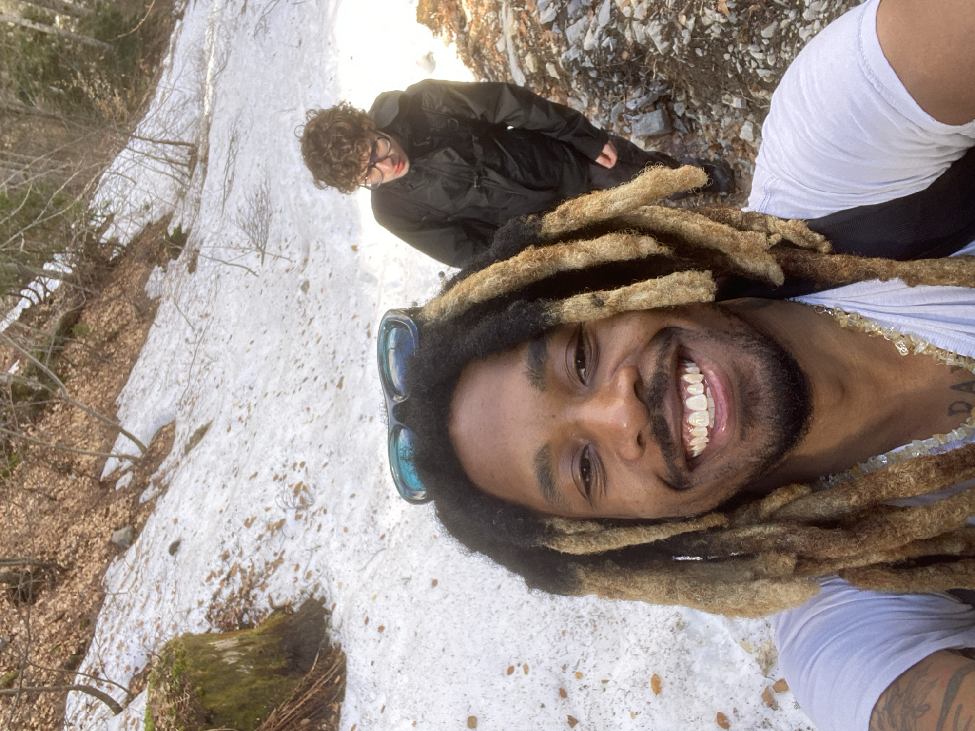 Two individuals are present in the image, taken outdoors in a snowy, mountainous environment potentially near Saint-Michel-les-Portes, France. One individual is positioned in the foreground, taking a selfie while smiling. They have dreadlocks, sunglasses resting on their head, and are wearing a white shirt. A beaded necklace is visible around their neck. The second individual is standing behind the first, on a rocky surface. They are wearing glasses and a black jacket. Snow is present on the ground and a forested area is partially visible in the background, including leafless trees and dead leaves.