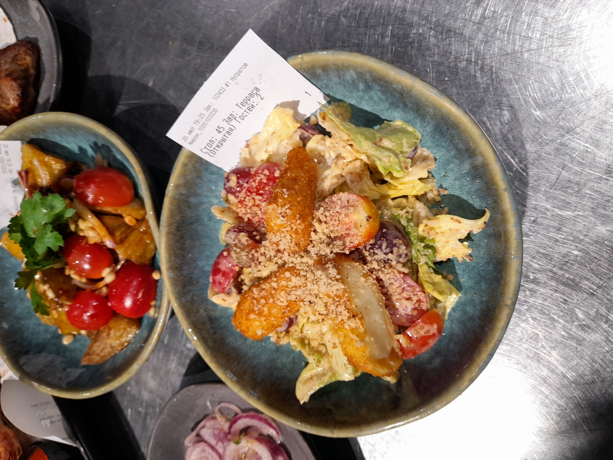 The image shows a close-up of a salad served in a teal-colored bowl, placed on a metallic, scratched surface. The salad contains lettuce, cherry tomatoes, possibly grapes, and pieces of fried cheese or halloumi, all coated in a creamy dressing and sprinkled with a nutty topping. A receipt is partially visible in the bowl, with Cyrillic text indicating a table number "45" and "Guests: 2". The receipt also contains a date and time: "20 июл 19:25".
The broader context, as indicated by the presence of a receipt with Russian text, suggests the location is likely a restaurant or cafe in Russia. The specific mention of "Терраса" (Terrace) on the receipt could imply an outdoor seating area. Without further visual cues or external information, pinpointing the exact establishment in Vladivostok is not possible from this image alone. However, given the details on the receipt, it's a plausible scenario for a restaurant in Vladivostok.