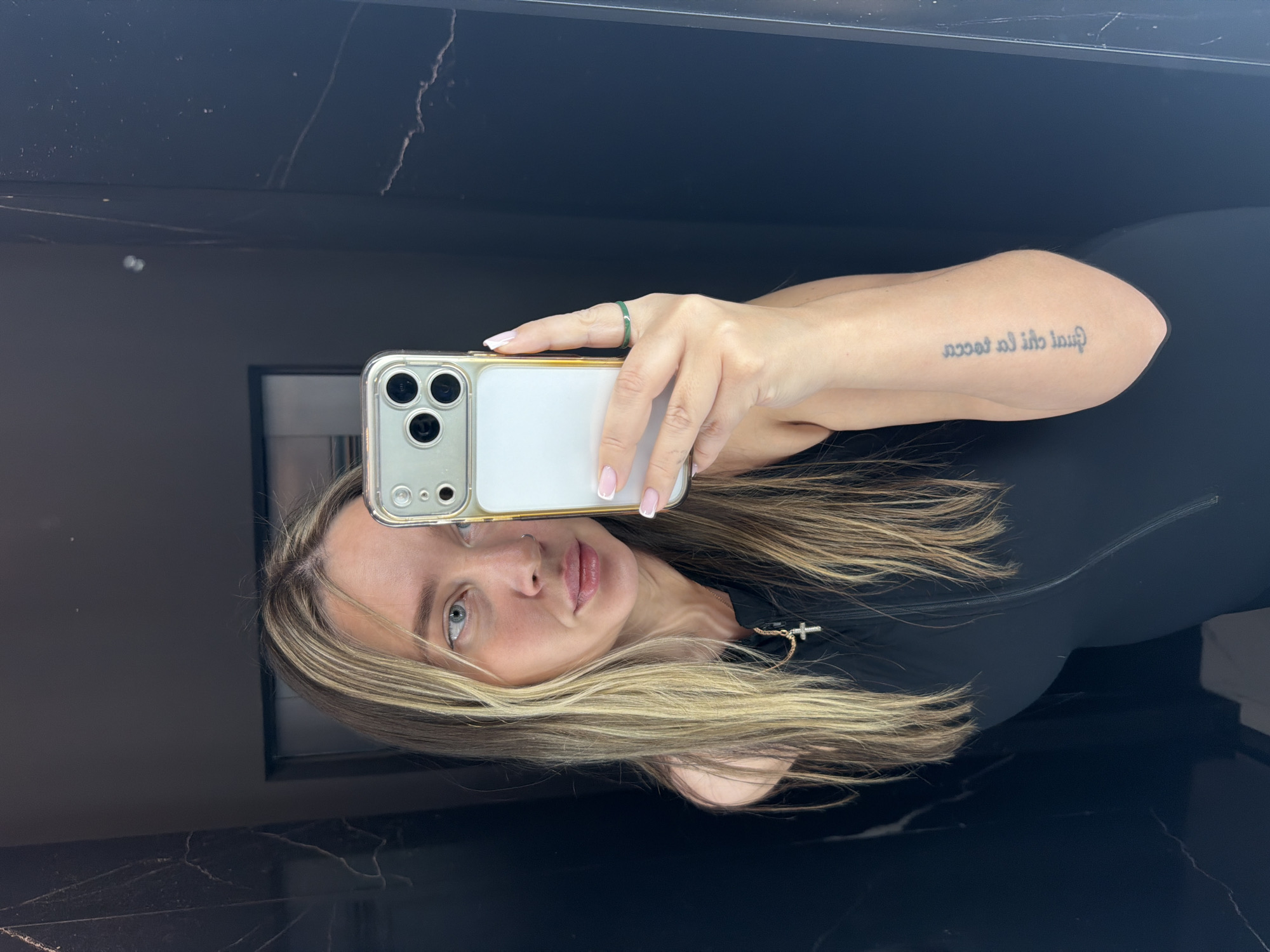A woman is taking a selfie in a bathroom. She is holding a white and gold iPhone in front of her face. She has blonde hair with dark roots and is wearing a black top with a small cross pendant. She has a ring on her right ring finger and a tattoo on her left forearm. The background is a dark wall, possibly made of marble or another shiny material, reflecting the light and the woman's image. There is a dark, rectangular object behind the woman, potentially a mirror or window. There is no indication of the city of Moscow, Russia, in the image.
