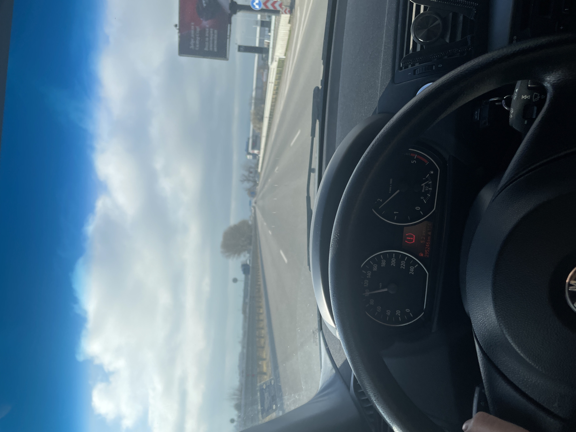 The image shows a driver's perspective from inside a car traveling on a highway. A billboard advertising "Judge amount to tasting cafe" is visible on the left. Other details include a clear blue sky, the road ahead, and the dashboard of the car. The speedometer indicates a speed around 100 km/h, with the engine at approximately 1500 RPM. The dashboard displays a warning light and information indicating 295,246 km, likely an odometer reading. Based on the billboard and landscape, the location is likely the Sofia ring road in Bulgaria.