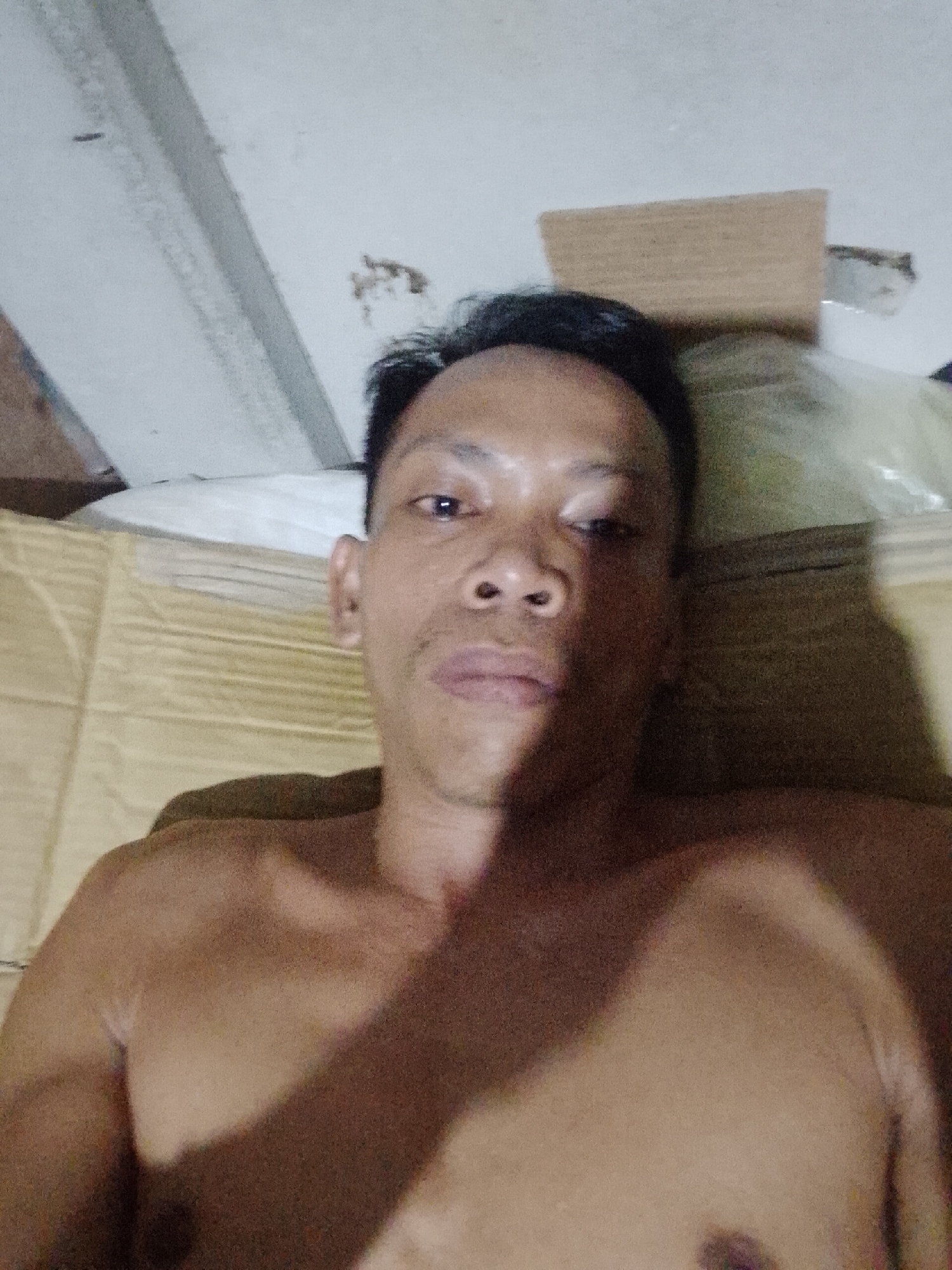 A medium shot photograph of an Asian man with dark hair and medium brown skin is lying on his back. He is shirtless, and his arms are visible. He is lying on what appears to be a mat or a soft surface with a repeating pattern. Behind his head is a white pillow and a cardboard box. The ceiling is visible at the top of the frame. The background has mold on the ceiling. The location is implied by the prompt.