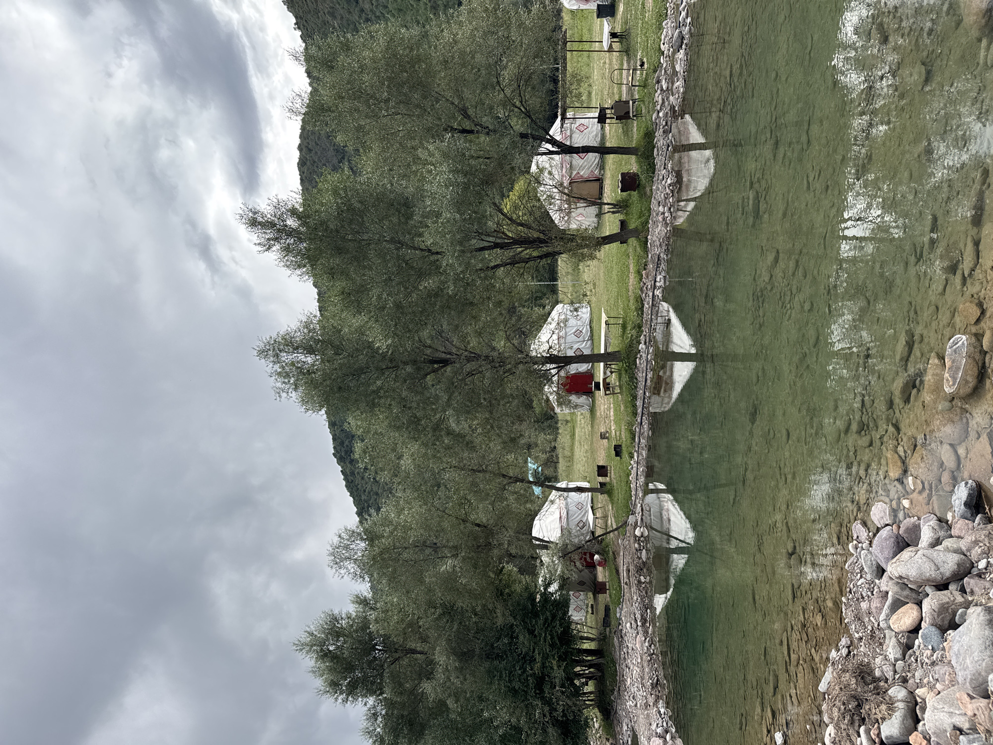 The image depicts a scenic view of a yurt camp situated alongside a clear, shallow river. The camp features several white yurts with red accents, set up on a grassy clearing amidst a line of lush green trees. In the background, a forested mountain slope rises under a cloudy, overcast sky. The river in the foreground is calm, with visible rocks on its bed and the reflections of the yurts and trees on its surface. The shoreline is rocky and natural. There are no people visible in the scene. The location is identified as Leskhoz, Kazakhstan.