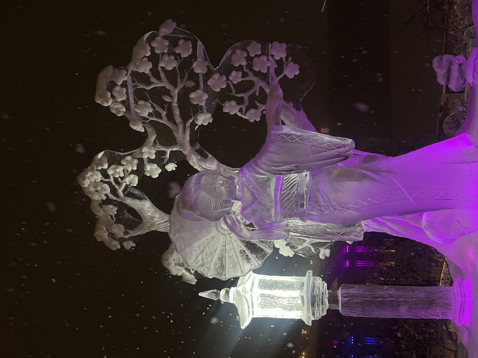 The scene depicts an outdoor ice sculpture display in Krasnoyarsk, Russia, at night during active snowfall. The primary subject is an ice sculpture of a woman, rendered in traditional Japanese attire, holding an open fan to her left. This figure is primarily illuminated from below by purple lights. To the woman's left stands an elaborate ice sculpture of a blossoming tree with multiple branches and distinct flowers, illuminated by white light. Adjacent to the woman and the tree, a vertical ice sculpture of a traditional Japanese-style lantern emits a bright white light. The base of the sculptures rests on accumulated snow, also accented by purple illumination. The sky is dark, consistent with nighttime conditions, and numerous snowflakes are suspended in the air. The distant background features indistinct, blurred lights, including purple and blue elements, indicative of an urban environment or further festive lighting.
