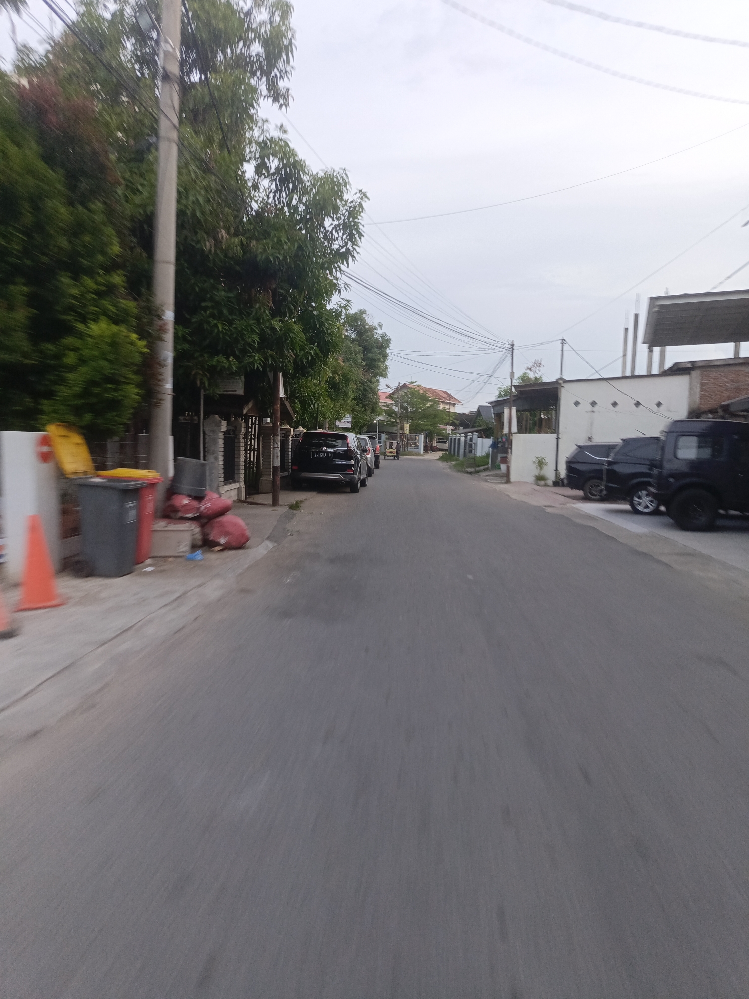 The image depicts a street scene in Lhokseumawe, Indonesia, under an overcast sky. On the left side of the road, a sidewalk is visible with a grey trash can topped by a yellow lid, and several large red trash bags. Further down the sidewalk, a red traffic cone is placed. Adjacent to the sidewalk is a line of trees and a utility pole with numerous wires. Several parked cars are seen along the right side of the road, including a black SUV and a black van. Buildings with various roofing styles are visible behind the cars and trees. The road itself is paved asphalt and appears empty in the foreground. The overall impression is a residential or commercial street on a cloudy day.