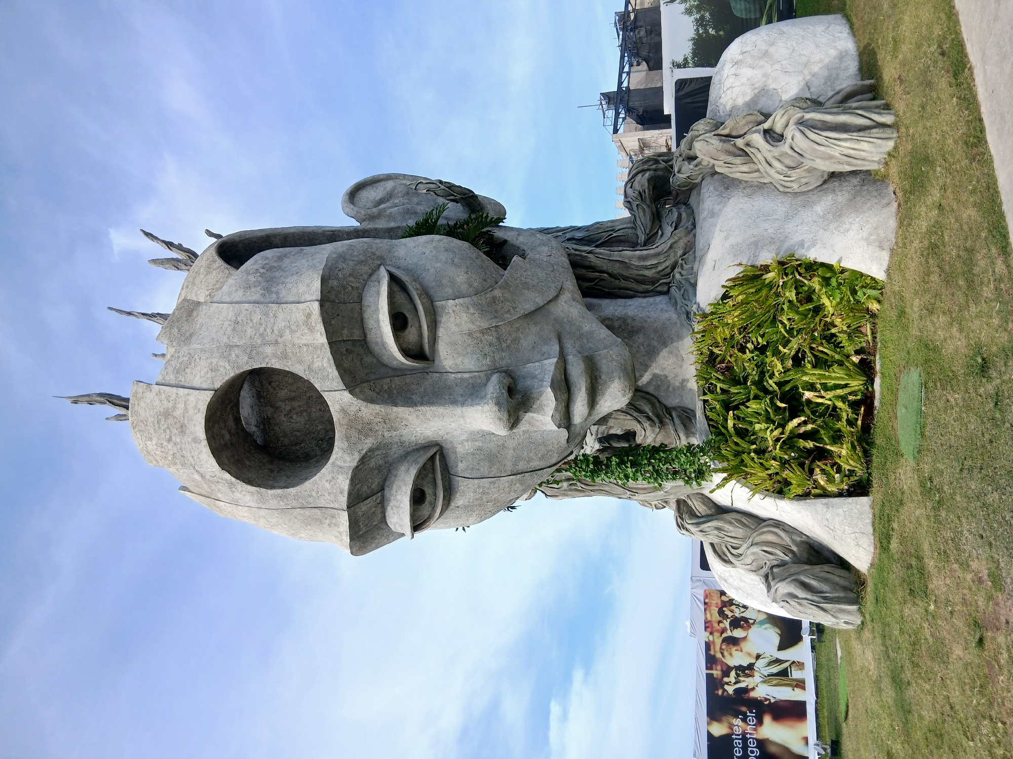 The image shows a large, imposing stone sculpture of a woman's head and upper torso situated outdoors on a grassy area. The sculpture has a somewhat abstract and stylized design, with a large circular opening in the forehead, elongated eyes, and braided hair that appears to be made of stone. Green foliage is integrated into the sculpture, growing from the chest area and within the ear. In the background, there are large banners, one displaying text and images of people, and other structures that appear to be part of an outdoor event or exhibition. The sky above is blue with scattered clouds. The sculpture's location is identified as Kediri, Indonesia.