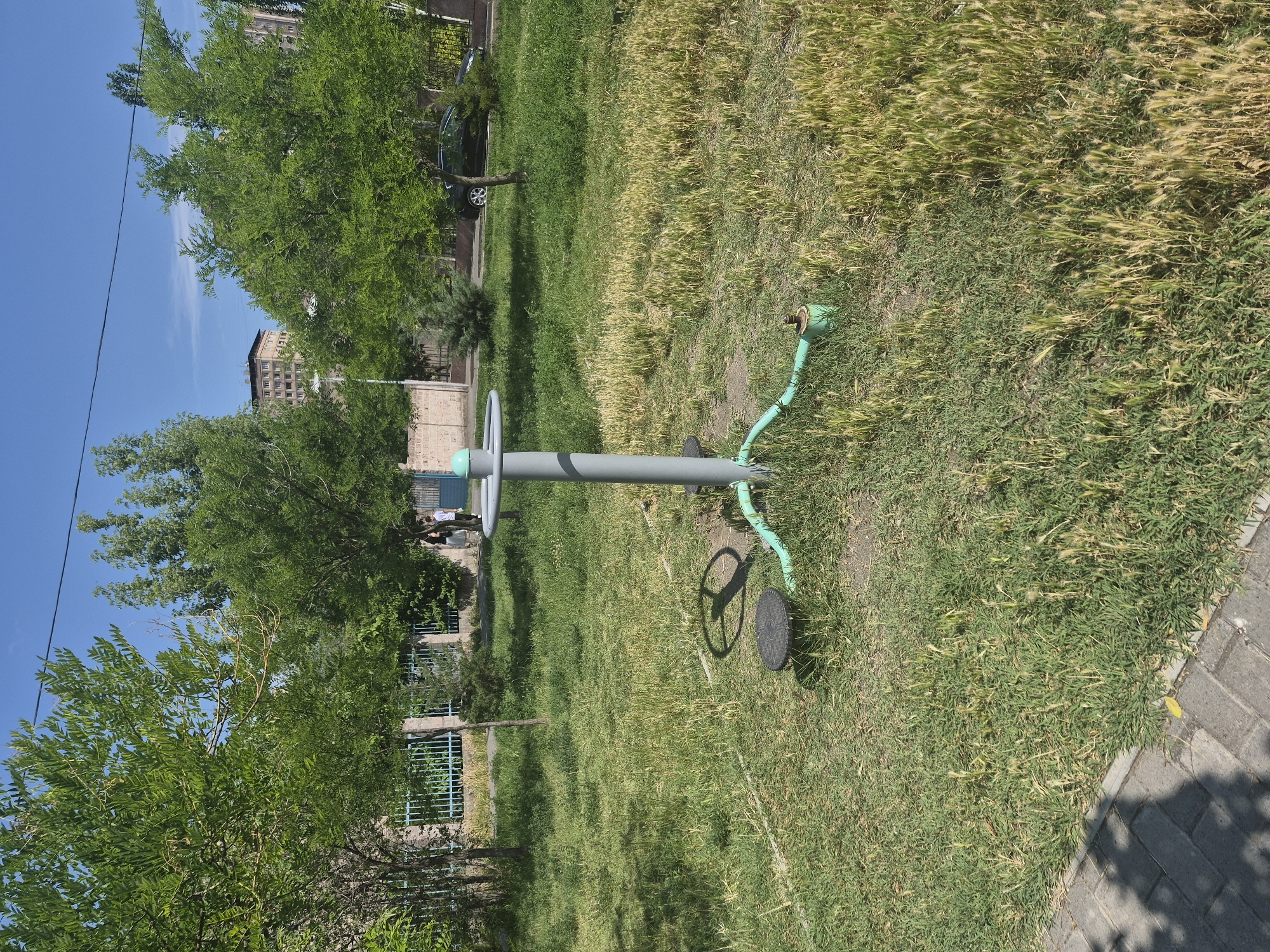 A gray metal outdoor fitness machine with a green base and turquoise cap is positioned in a grassy park area. The machine has a central pole and two circular footrests. A shadow of the machine, cast by the sun, is visible on the grass. The park is bordered by a light-colored paved walkway on the left and a light-colored brick wall with a blue fence on the right. Mature trees with green foliage are present throughout the scene. In the background, two people, one wearing a dark top and the other a white t-shirt, are walking away from the camera. A dark-colored car is parked on a street behind the fence, and apartment buildings are visible further in the distance. The sky is clear and blue. The location is the Abovyan Park in Yerevan, Armenia.
