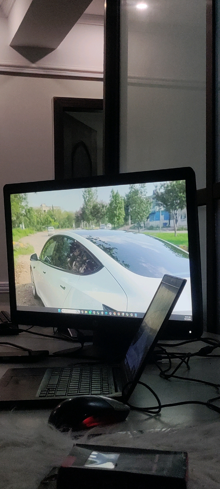 The image depicts a desk setup with a large monitor displaying a white car on a road with trees. In front of the monitor, a laptop is open and angled towards the viewer, with its screen partially visible. A black computer mouse with a red illuminated scroll wheel is positioned on a white, fuzzy surface in front of the laptop. Cables are visible connected to the devices. The background shows a dimly lit room with a ceiling fan and a doorway with a dark frame. There are no people or animals visible in the image. The location in the city of Khana Dak, Pakistan, is not discernible from the visual information provided.