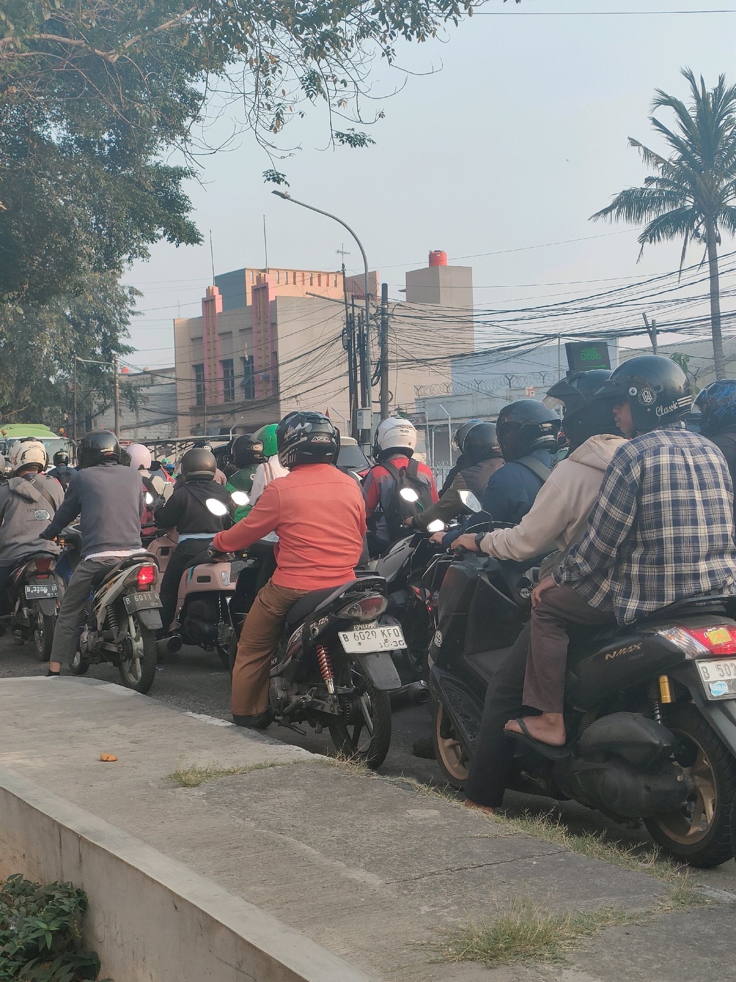 The image depicts a congested street scene with a line of motorcycles and riders, likely in the city of Bekasi, Indonesia, given the context. The time appears to be late afternoon or early evening, as indicated by the warm lighting and long shadows.
In the foreground, several motorcycles are stopped in traffic. Riders are wearing helmets, and some are looking ahead, while others appear to be in conversation or simply waiting. The motorcycles are of various models, with some displaying license plates.
In the mid-ground, more motorcycles and riders are visible, further emphasizing the traffic situation. The background reveals urban infrastructure, including buildings with distinctive architecture, utility poles with a dense network of wires, and street lights. A tall palm tree can be seen on the right side of the frame. The overall atmosphere suggests a typical urban commute in a densely populated area.