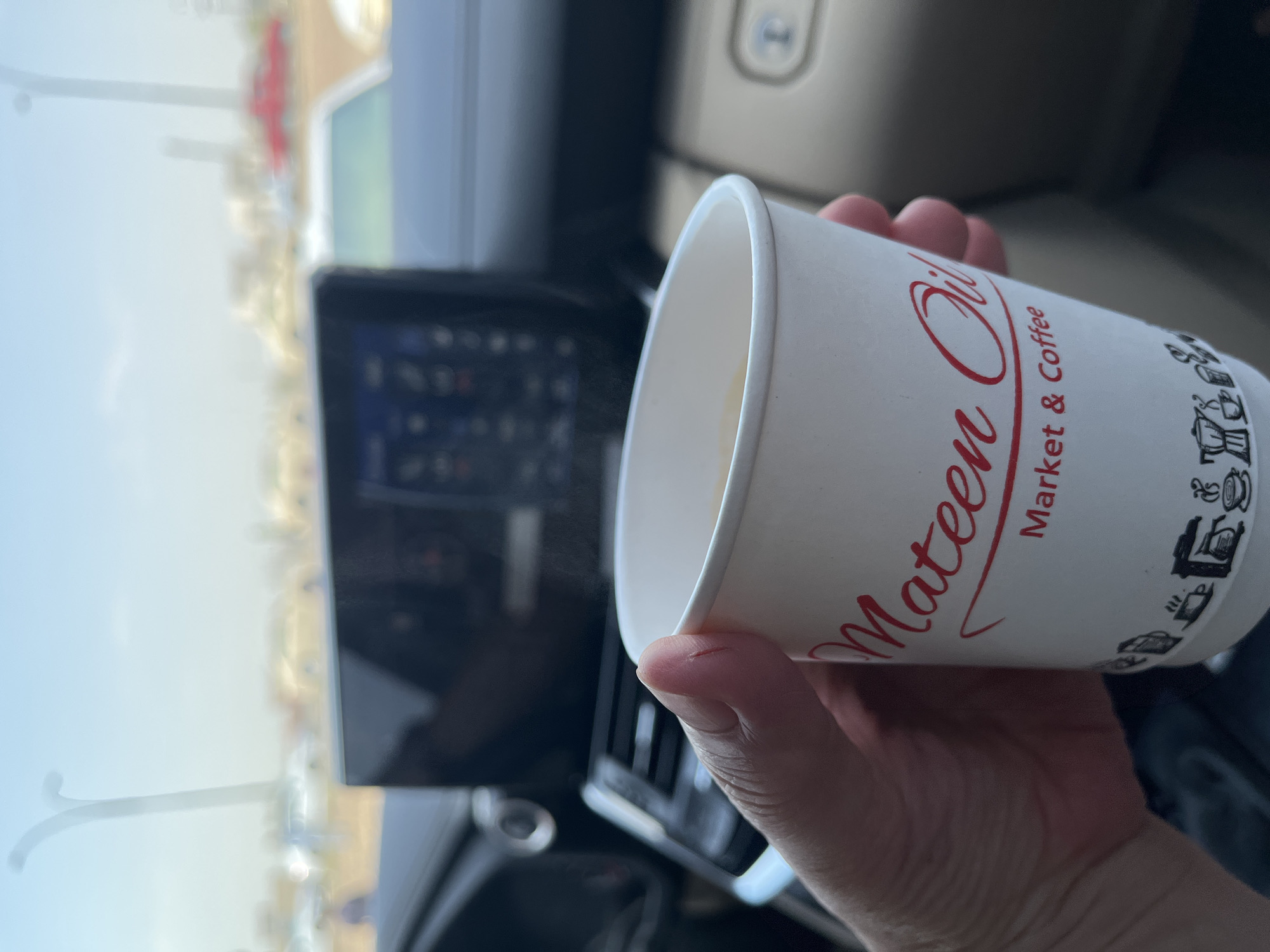 A close-up shot shows a hand holding a white paper coffee cup with red text that reads "Mateen Oil Market & Coffee." The cup is partially filled with a light-colored beverage, and a faint wisp of steam rises from it. The background is out of focus, revealing the interior of a vehicle, including a dark dashboard with a digital display and a portion of the steering wheel. Beyond the car's windows, a blurry urban landscape is visible under a pale sky, with hints of cars and streetlights.