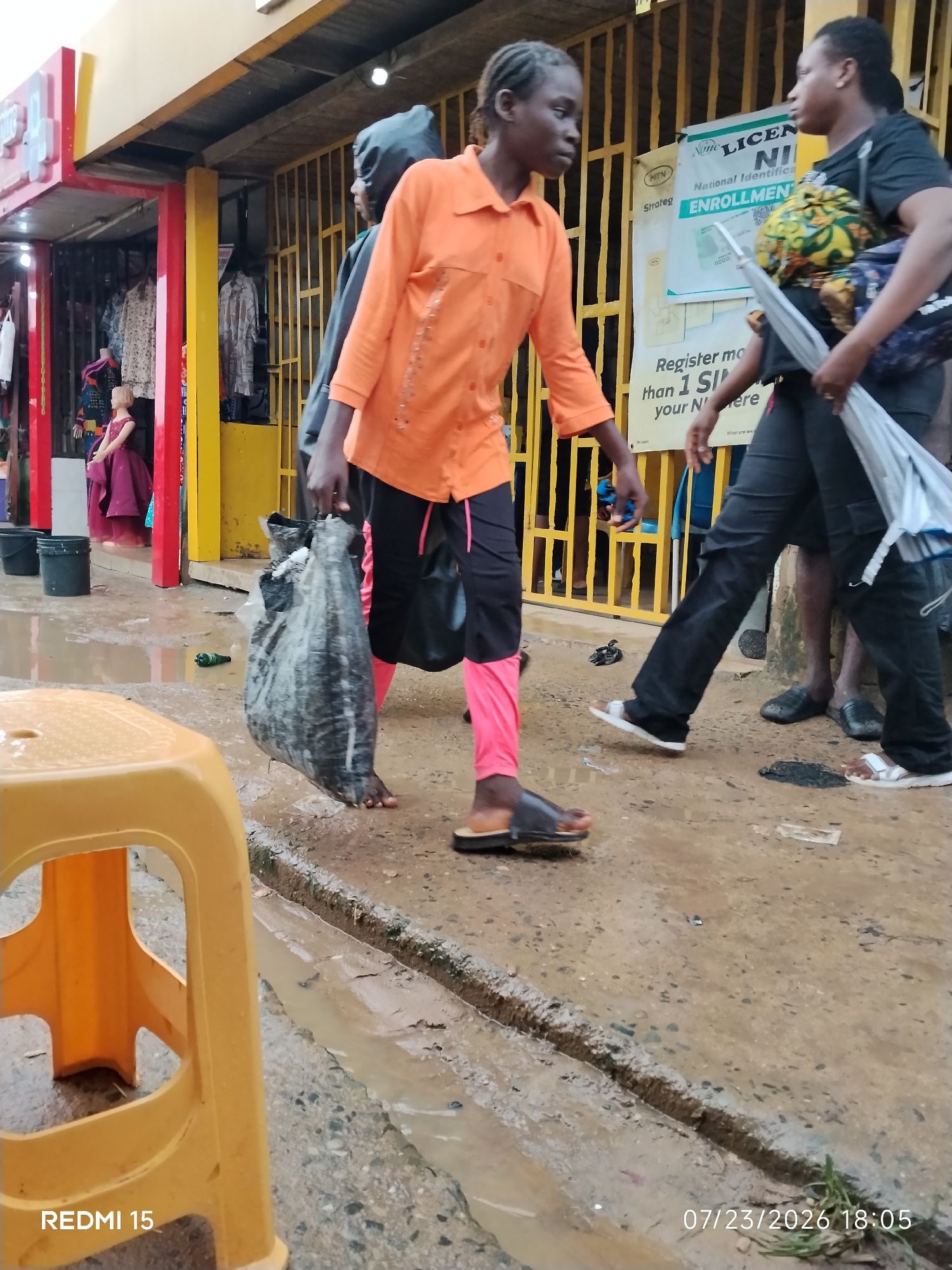 In Umuode, Nigeria, a young Black woman in an orange shirt and pink pants walks across a wet, muddy street, carrying a large, dark plastic bag. She is barefoot and wearing dark sandals. The street is lined with shops and stalls, with yellow bars visible on some structures. A yellow plastic stool is in the foreground to the left, and a poster with text in English is mounted on a yellow grate behind the woman. The environment appears to be a marketplace or commercial area, with puddles of water on the ground indicating recent rain.