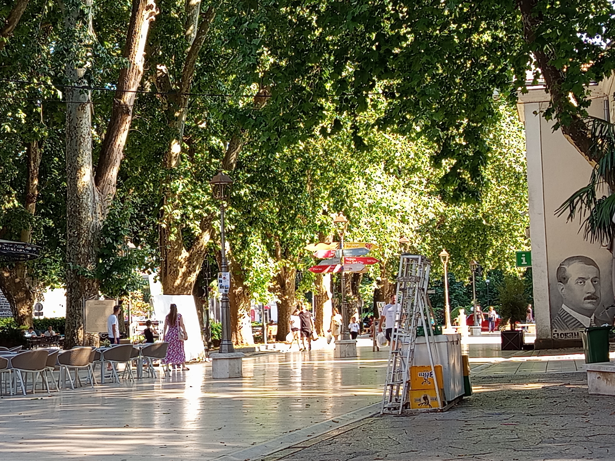 The image shows a public square in Trebinje, Bosnia and Herzegovina, on a sunny day. A wide, paved walkway is lined with large trees, casting dappled shadows. On the left side of the walkway, outdoor seating areas with tables and chairs are visible. In the mid-ground, several people are walking or standing. A woman in a floral dress is walking away from the camera, carrying a bag. Further down the walkway, a man in a white t-shirt and shorts is walking towards the camera. On the right side of the frame, a tall metal ladder stands next to some stacked yellow crates. A mural of a man's portrait is painted on a building wall to the right, with the name "JOVAN" written below it. A green trash can is also present on the right. The atmosphere appears relaxed, suggesting a leisurely day in the city.