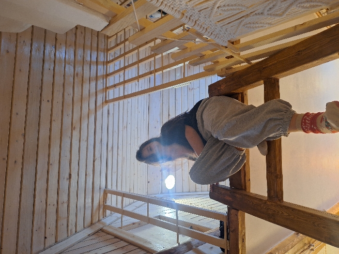 The scene depicts an interior space within a building in Lezhnevo, Russia, characterized by extensive light-colored wooden construction on the walls and ceiling. A female individual is positioned on a rustic wooden ladder, seated on a horizontal rung with her legs bent. She wears a dark top, loose grey sweatpants with elasticated ankles, a white sock on her right foot, and a red sock with white patterns on her left foot, which rests on a lower rung. Her dark hair is visible, and she looks upwards towards the camera with a slight smile. A bright light source behind her head creates glare, partially obscuring her face. The ceiling above consists of closely spaced wooden planks. Vertical wooden beams serve as structural elements and form a partial railing for an upper level. A white macrame decoration is partially visible on the far right. The image is captured from a low-angle perspective, looking upwards at the individual and the surrounding wooden architecture.