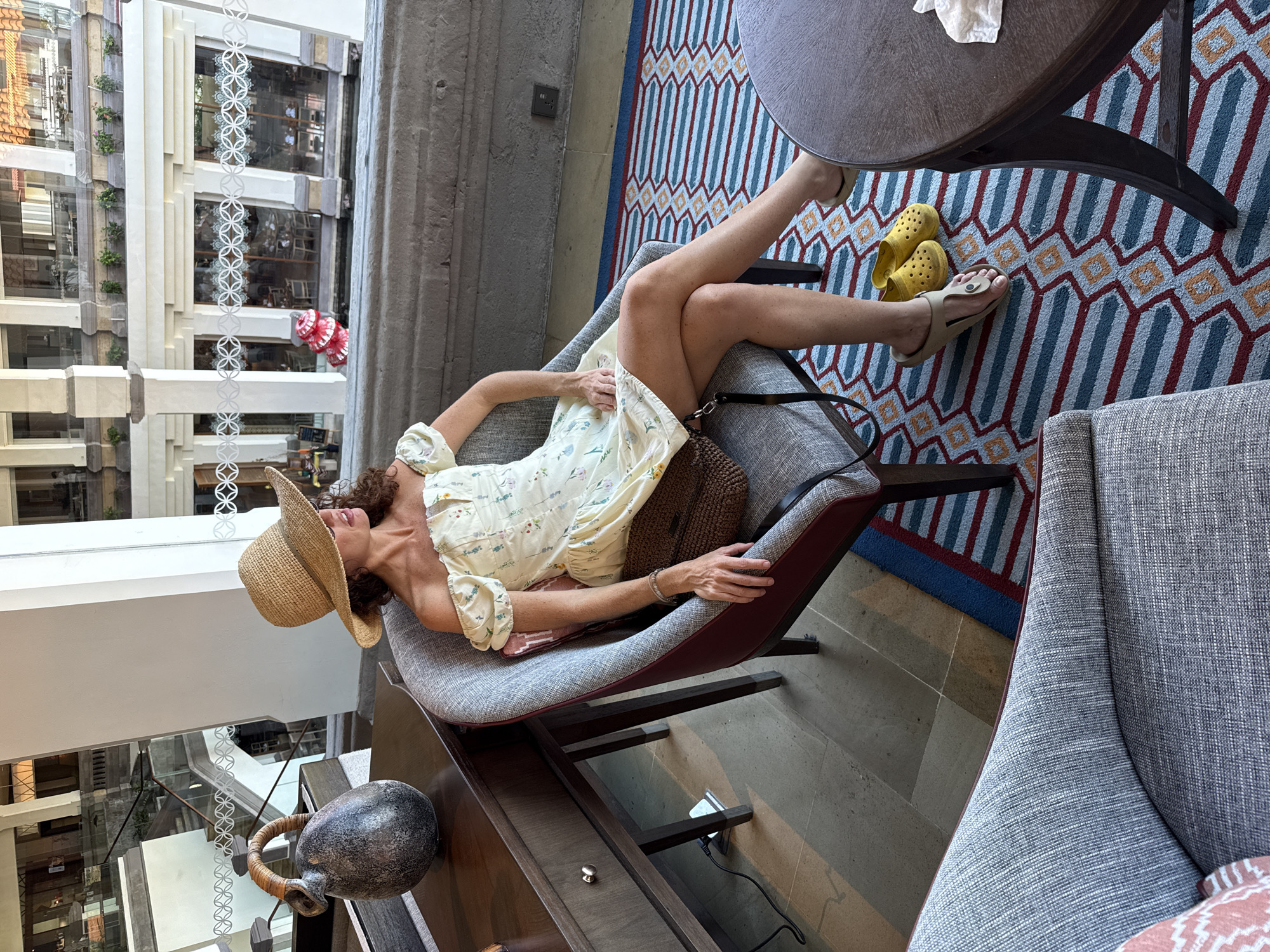 A woman wearing a straw hat and a yellow off-the-shoulder dress with a floral pattern sits in a grey armchair on a colorful geometric rug. She has a brown bag on her lap and is wearing tan flip-flops and yellow Crocs on her feet. She is looking to her right. Beside her, to her left, is a dark wood desk with a grey ceramic vase on it. In the background, a multi-story interior with white columns and windows is visible. The scene appears to be a hotel lobby or common area in Kuta Selatan, Indonesia.