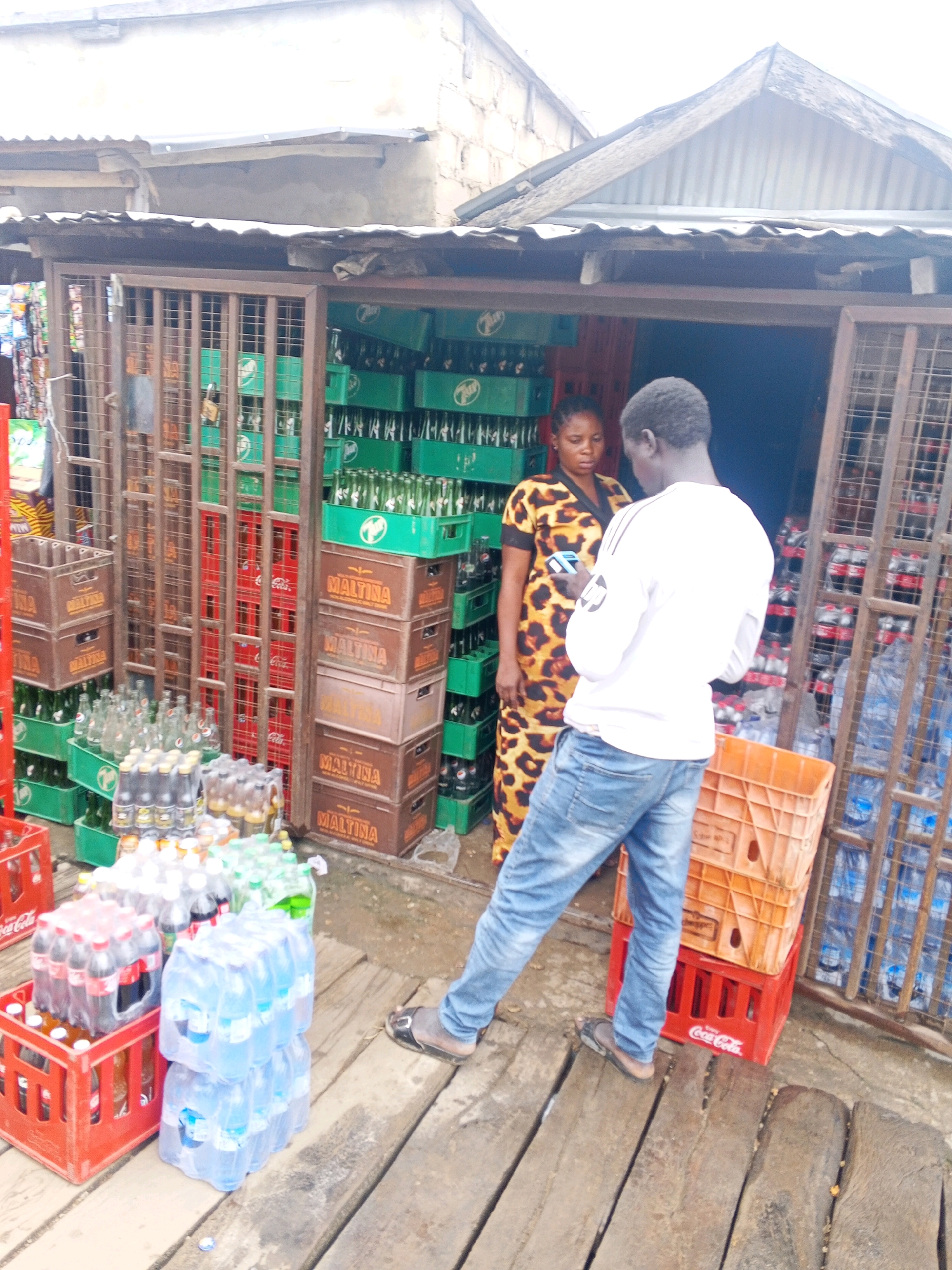 The image depicts an outdoor roadside retail stall in Lokoja, Nigeria, characterized by a structure with a corrugated metal roof and a facade of metal bars. The stall is laden with a variety of beverages, primarily bottled water and soft drinks, displayed in crates and on shelves. Stacks of green crates containing bottles are visible, along with brown crates labeled "MALTINA." Various bottles of water are also present, some in plastic packaging.
In the foreground, wooden planks form a makeshift walkway in front of the stall. Two individuals are interacting at the entrance of the stall. A man wearing a white long-sleeved shirt and blue jeans is facing away from the camera, looking down at his mobile phone. He is wearing sandals. A woman, standing next to him and facing the stall, is wearing a leopard-print dress. She is also looking at her phone. The overall environment suggests a busy, informal commercial setting typical of Nigerian street markets.
