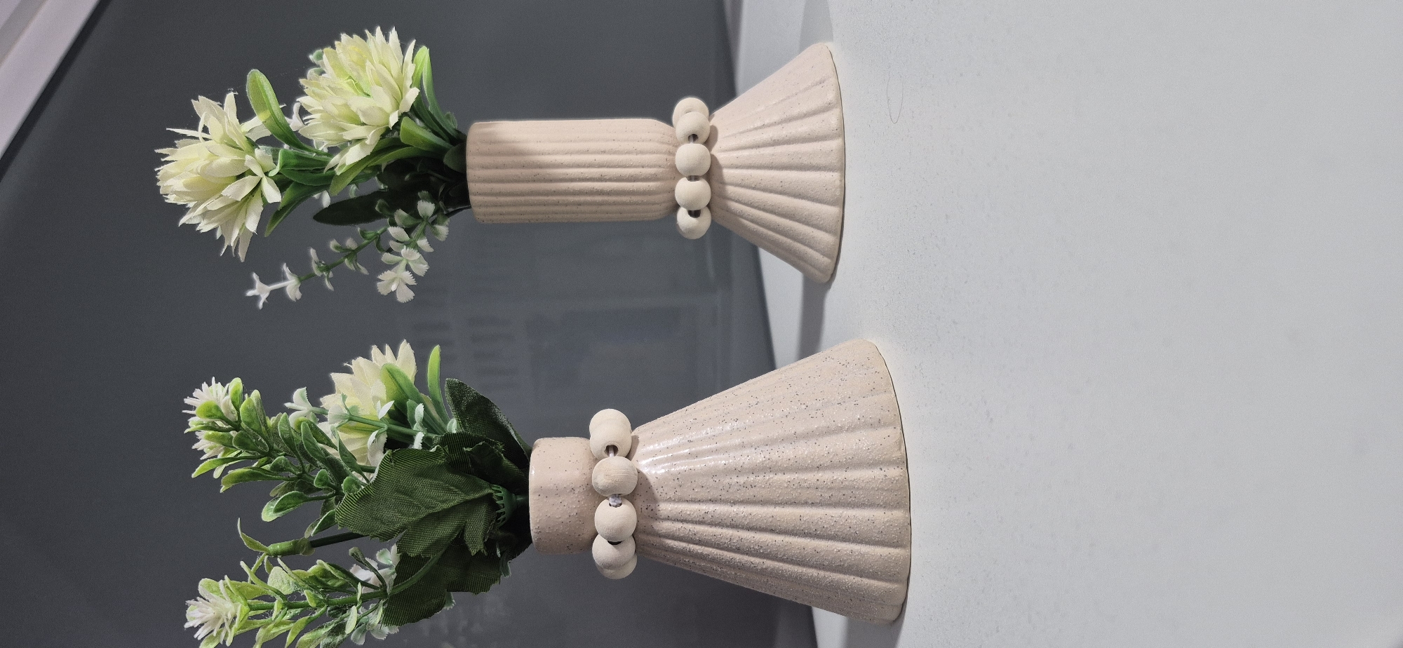 The image displays two beige ceramic vases with ribbed textures and decorative beaded accents. Both vases contain artificial flowers and greenery. The vase on the left has a flared, cone-like base, while the vase on the right has a cylindrical upper section and a flared base. They are placed on a white surface against a dark gray background. The scene is evenly lit, casting soft shadows. The image does not contain any information about the city of Vila do Conde, Portugal.