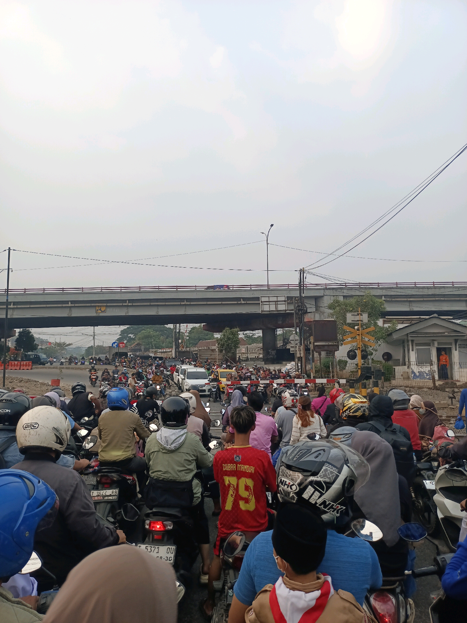 The image captures a congested traffic scene in Karawang, Indonesia, likely near a railway crossing. A multitude of motorcycles and a few cars are stalled on a road under an elevated overpass. People on motorcycles are mostly wearing helmets, with various colors and designs visible. Some individuals are wearing masks. The environment appears to be a city with a clear sky, overhead power lines, and a railway crossing signal with barrier gates. Buildings and trees are visible in the background, suggesting an urban or semi-urban setting. The overall impression is one of a typical traffic jam in a bustling Indonesian city.