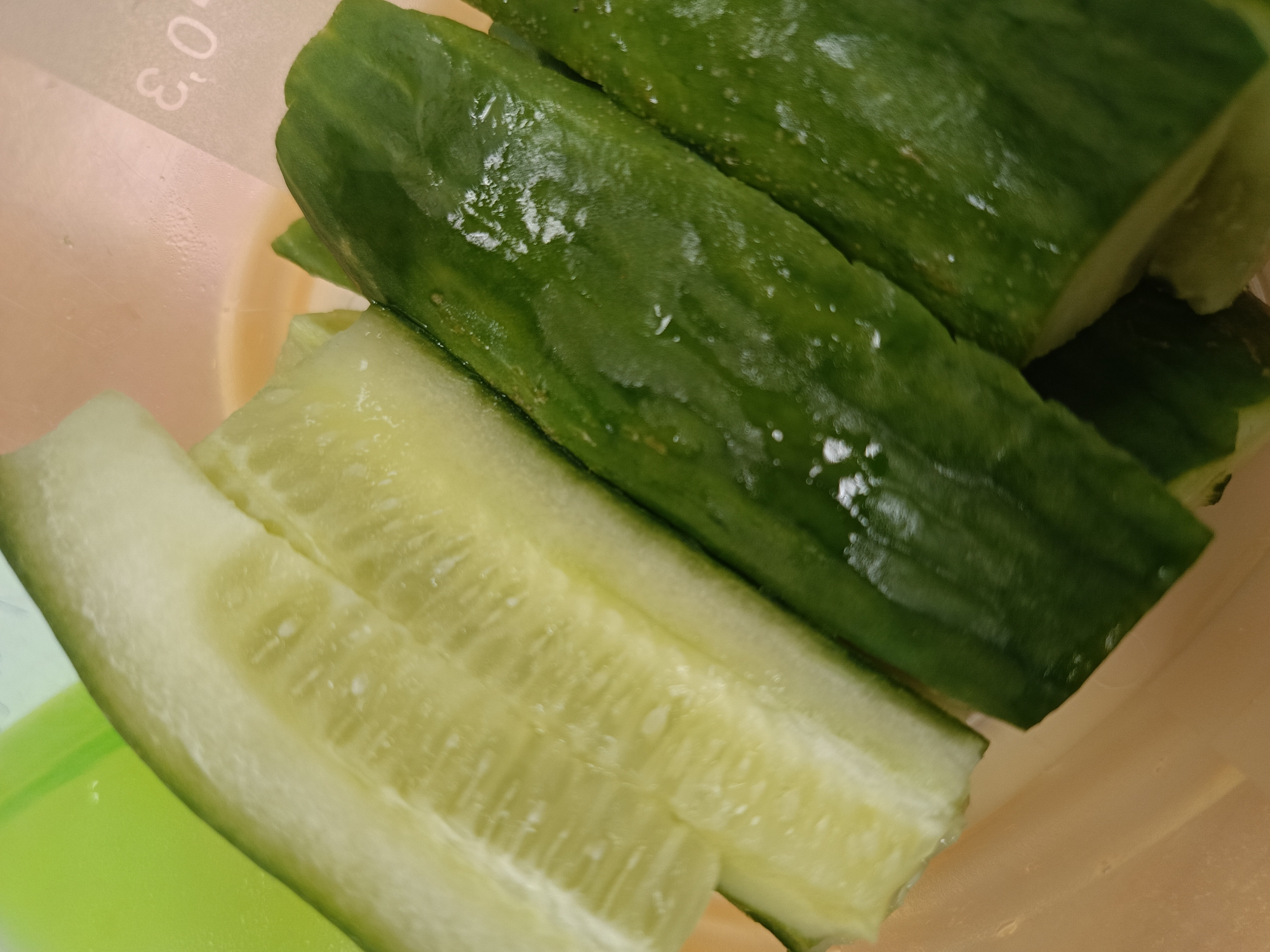 The scene captures a close-up, high-angle view of freshly cut cucumber pieces contained within a plastic vessel. One prominent piece is longitudinally halved, displaying its pale green flesh, visible seed cavities, and internal texture. Several other dark green cucumber segments, still retaining their textured skin and glistening with water droplets, are arranged around it. The cucumbers are situated within a container featuring at least two plastic components: a translucent, light beige or pale orange section with faint embossed markings '0.3' visible, and a distinct bright lime green section in the lower left portion of the frame. No individuals, actions, or specific interactions are depicted. The image does not contain visual information to ascertain its location within the city of Chaikovsky, Russia, beyond the immediate container and its contents.