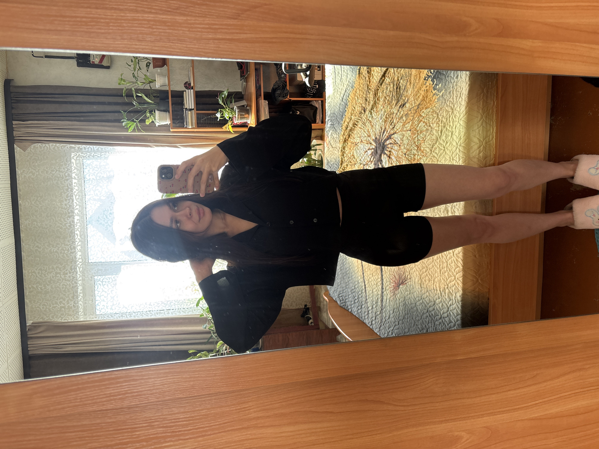 The image is a mirror selfie of a woman with long dark hair. She is wearing a black button-down shirt and black shorts, and pink slippers. She is holding a cell phone in her hand. Behind her, the reflection in the mirror shows a window with sheer curtains, a shelf with plants, and a bed with a patterned comforter. The mirror is framed with a wooden border. The location is an indoor residential setting, but there is no indication of the city of Dubrova, Russia in the image.