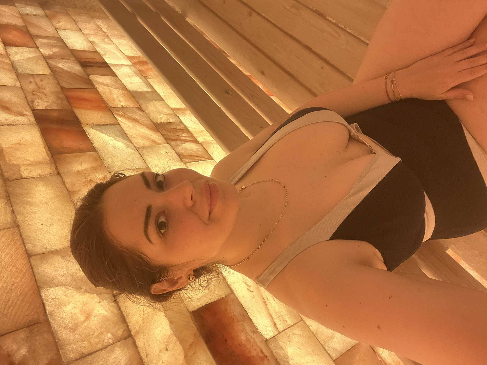 A woman is lying down in what appears to be a sauna, with a floor constructed of illuminated salt bricks. The sauna benches are made of light-colored wood. The woman is wearing a dark-colored, possibly black, garment with light-colored straps. She has light skin and dark hair. She is wearing a necklace and bracelets. It is not possible to determine the specific location in Kazanlak, Bulgaria from the image alone.