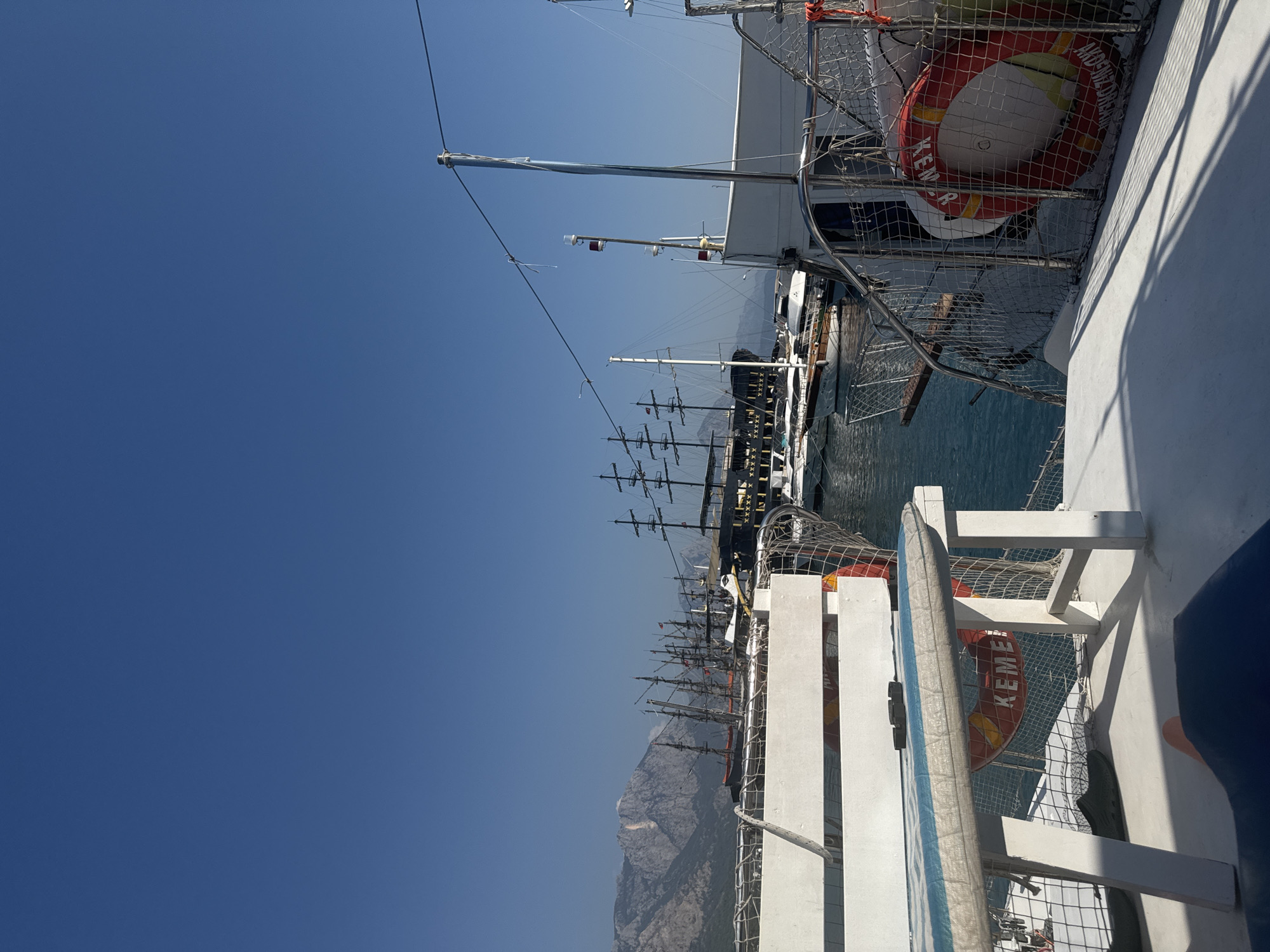 The image shows a view from a boat in the harbor of Kemer, Turkey. In the foreground, there is a white bench with a blue and white striped cushion. Behind the bench, there is a life preserver with the word "KEMER" visible. Several boats are docked in the harbor, including large sailing ships with multiple masts. In the background, there are mountains. The sky is clear and blue.