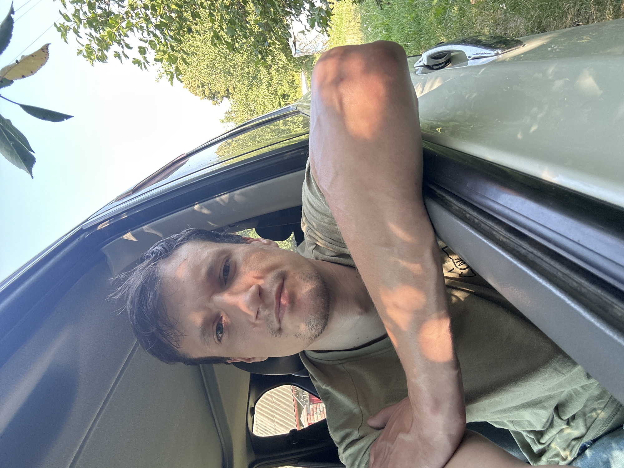 A young man with dark, tousled hair and a light stubble on his chin is pictured from the chest up, looking directly at the camera from inside a light-colored vehicle. He is wearing a plain olive-green t-shirt. His left arm is resting on the open car door, with his hand tucked behind his elbow. Sunlight casts a warm glow across his skin, particularly on his left arm and upper chest. The background shows greenery and trees, suggesting an outdoor setting. A portion of a building with a red-tiled roof is visible through the rear passenger window. The overall scene appears to be in the city of Shatava, Ukraine.
