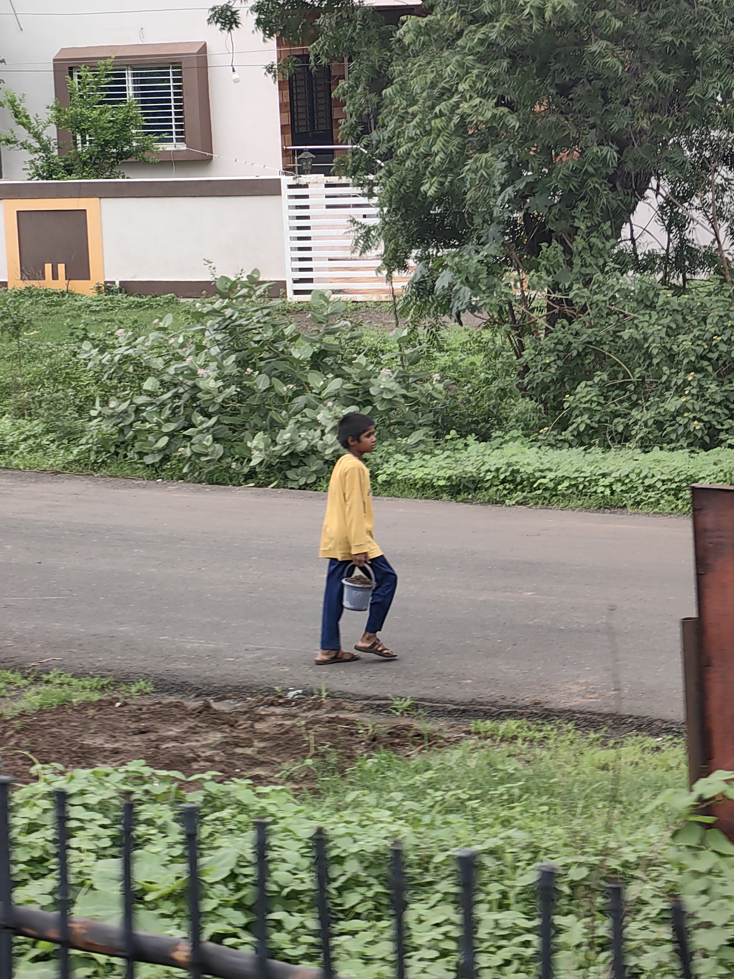 A young boy walks on a paved road in Sakari, India, carrying a light blue bucket in his right hand. He is wearing a yellow long-sleeved shirt and dark blue pants. The road is flanked by lush green vegetation and some trees. In the background, there is a white building with brown trim and a window with a security grate. A black metal fence is visible in the foreground.