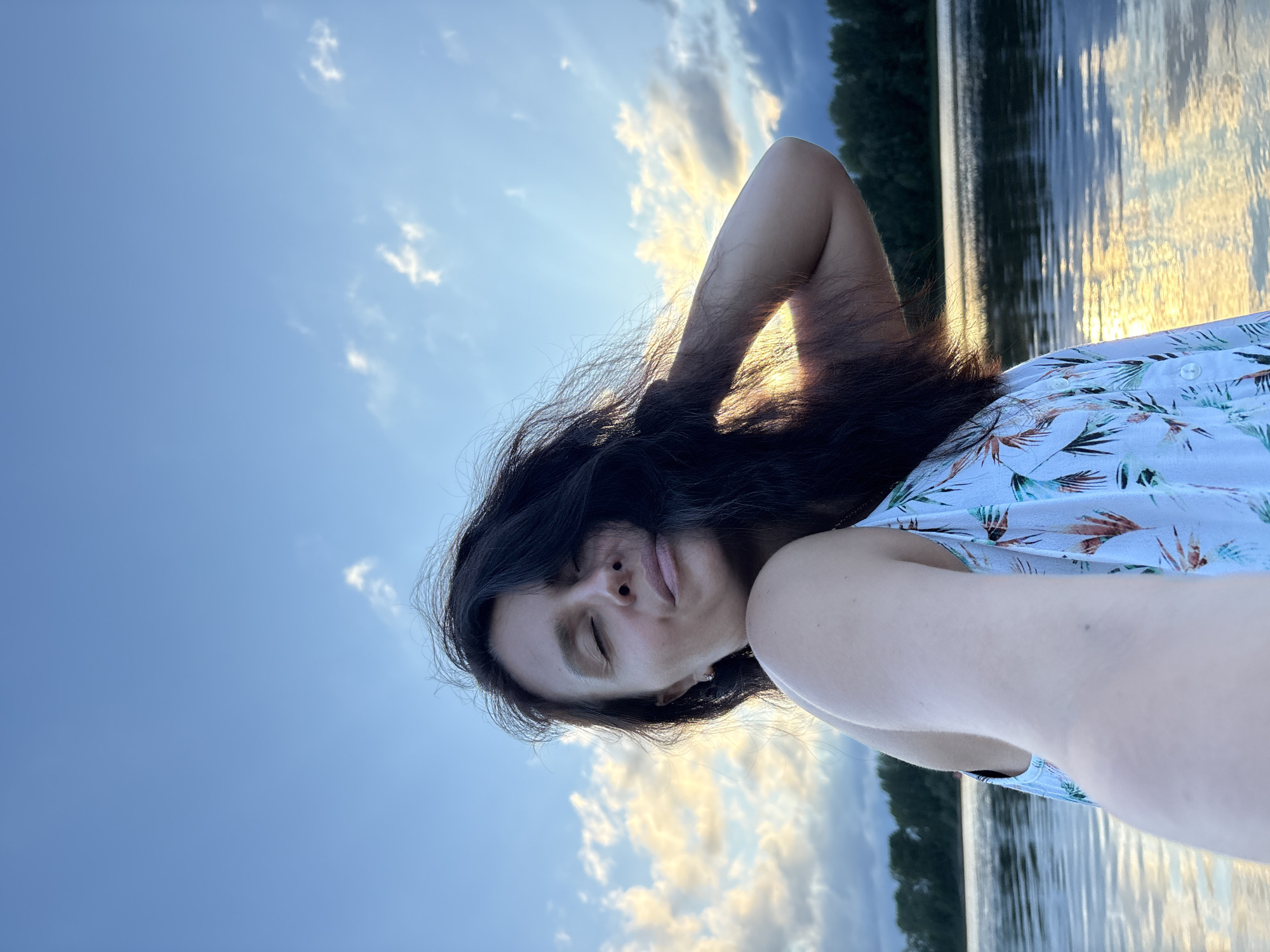 A woman is taking a selfie outdoors by a body of water. The woman has dark hair and is wearing a white dress with a tropical pattern. Her eyes are closed, and she is touching her hair with her right hand. The sky is blue with clouds and the sun is setting, casting a golden reflection on the water. In the background, there is a treeline. The location is the Kama River in Chaikovsky, Russia.