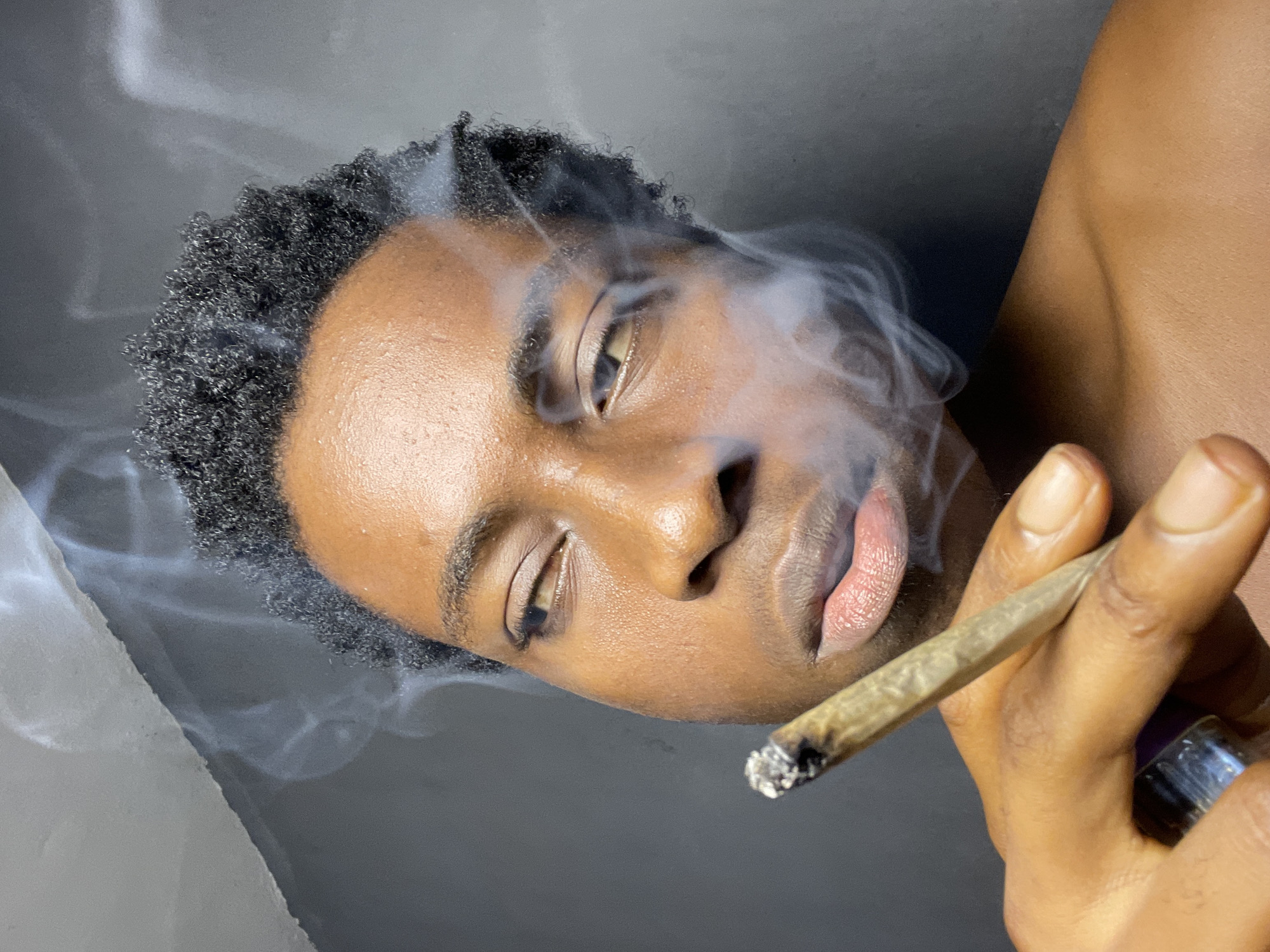A close-up, vertically oriented photograph depicts a young Black man with dark, curly hair and brown skin, smoking a marijuana cigarette (joint). He appears to be in a relaxed state, with a slight downward gaze. Smoke billows around his face. The joint is held in his right hand, and a purple/silver object is visible behind his fingers. The background is a neutral gray/white gradient. The location, Nkpor, Nigeria, is not evident within the image.