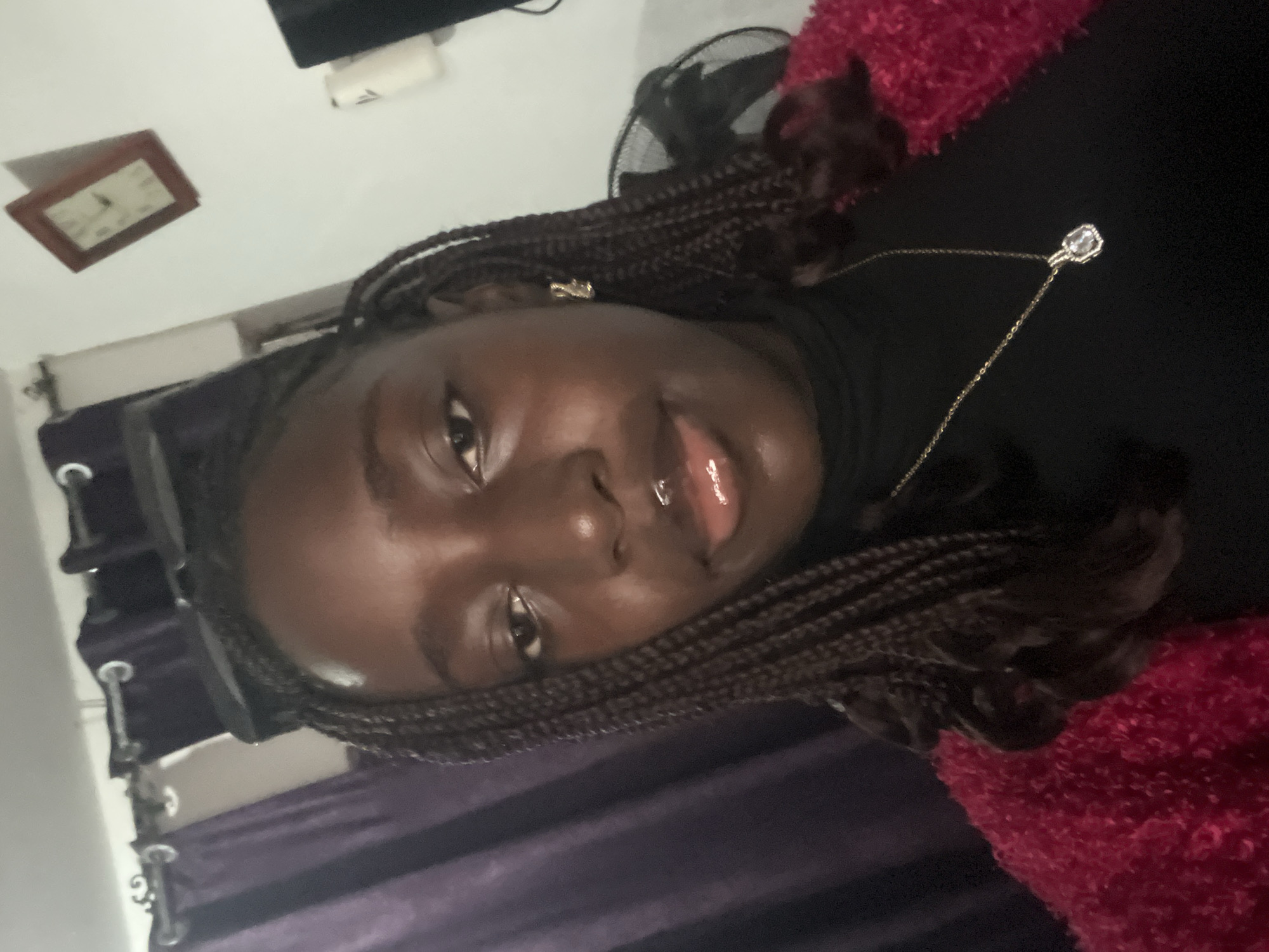 A person with dark skin and braided hair is in the foreground, wearing a black turtleneck shirt and a red fuzzy garment. The person has a gold necklace with a silver pendant. In the background, there are purple curtains, a clock on the wall, a white object, and a fan. The location appears to be an indoor setting. The city of Abuja, Nigeria is not discernible from the provided image.