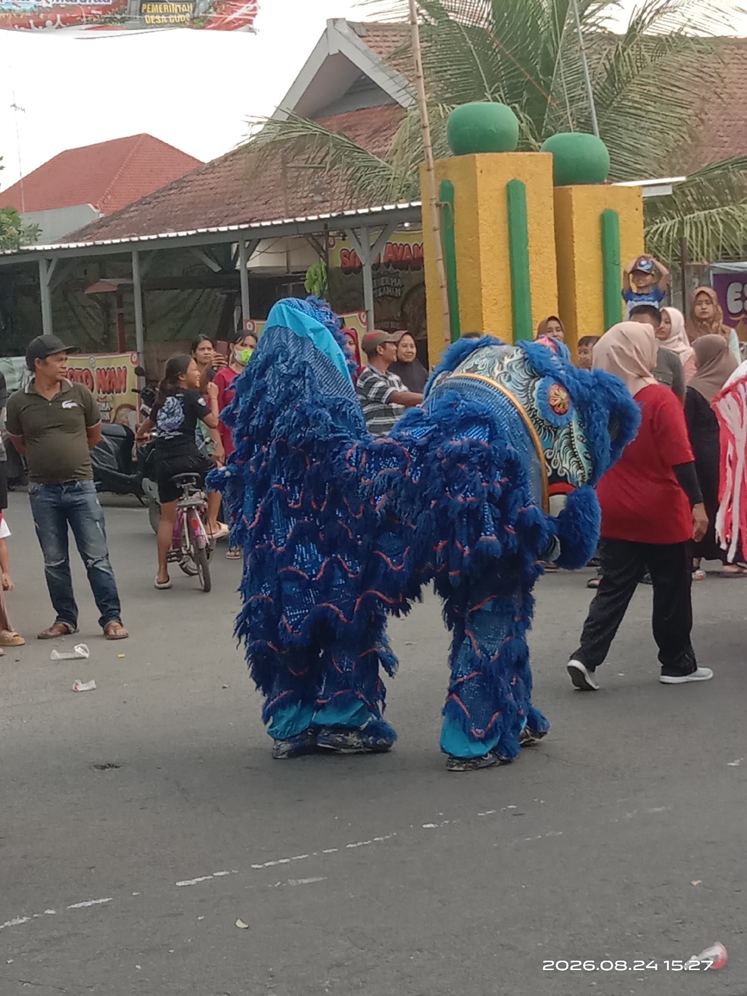 In Gudo, Indonesia, a lively street scene unfolds with a vibrant lion dance performance. The focal point is a large, blue lion costume, likely being operated by two individuals unseen beneath it, with its head adorned with ornate patterns and fluffy blue fur. The lion is positioned on an asphalt road, which appears somewhat worn.
Several people are gathered around, observing the spectacle. To the left, a man in a dark green polo shirt and distressed blue jeans stands watching, his hands in his pockets. Beside him, a woman rides a pink bicycle, also looking towards the lion. Further in the background, a crowd of onlookers, including women in hijabs, are visible.
Behind the crowd, buildings with red tiled roofs and white-trimmed eaves suggest a local urban environment. A decorative structure, possibly part of a celebration or event, stands tall with yellow and green pillars topped by green spheres. In the distance, palm trees indicate a tropical climate. The overall atmosphere appears festive and communal.