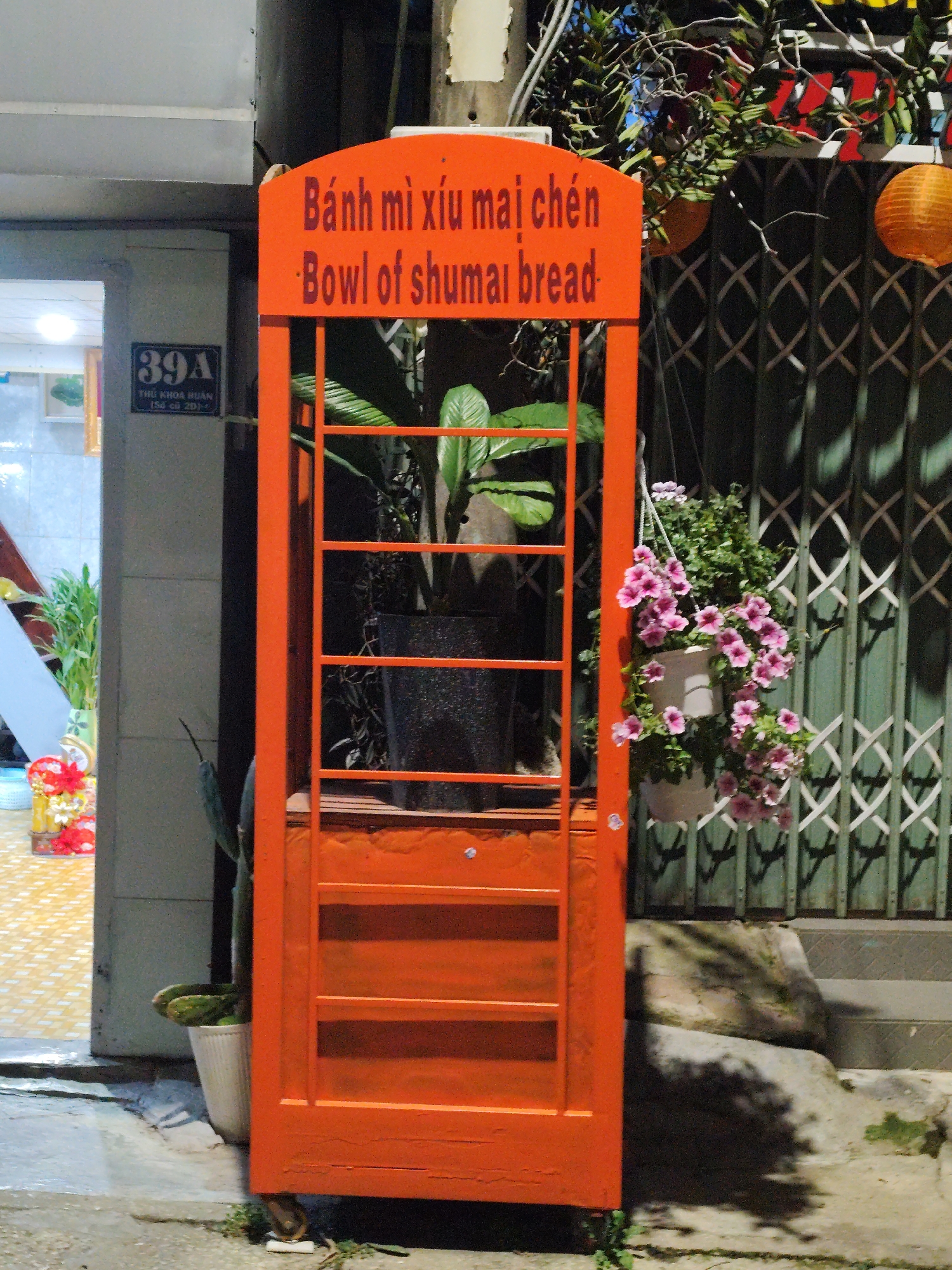 This media file depicts an outdoor night scene in Da Lat, Vietnam, focusing on a vibrant orange food stand or sign. The stand is tall and narrow, resembling a decorative phone booth with internal shelves or openings, and is made of wood painted in a bright, almost fluorescent orange.
At the top of the orange stand, text is visible in both Vietnamese and English. The Vietnamese reads "Bánh mì xíu mại chén," and the English translation below it states "Bowl of shumai bread," indicating that the stand likely advertises or sells this specific Vietnamese dish.
Inside the orange structure, a potted green plant is visible. To its right, a white hanging planter overflows with cascades of pink, petunia-like flowers. Behind these flowers and the stand, a dark green metal rolling gate with a diamond pattern is seen. Further to the right, an orange paper lantern hangs from above.
To the left of the orange stand, the entrance to a building is brightly lit, revealing a light-colored tiled floor inside and some decorative items. On the building's exterior wall, a blue address plaque is affixed, displaying "39A THỦ KHOA HUÂN (Số cũ 20)." A slender, cactus-like plant in a white pot stands at the base of the orange structure. The ground is concrete, and some rough stone steps are visible to the right of the stand. The lighting suggests it is nighttime, with artificial light illuminating the scene. No people are present.