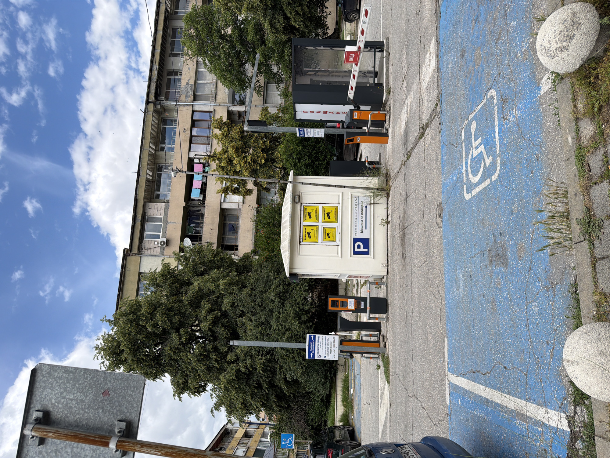 The image depicts an outdoor parking lot in Pazardzhik, Bulgaria. In the foreground, a blue-painted parking space with a white wheelchair symbol is visible. A large tree with dense green foliage dominates the mid-ground, partially obscuring a multi-story apartment building. Adjacent to the tree is a small, white booth with yellow signs displaying camera icons and text in Cyrillic. To the right of the booth, a parking barrier arm is raised, allowing entry into the parking area. An exit gate with a red and white striped barrier is also present on the far right. Several parking payment machines and traffic control equipment are situated near the booth. The sky above is blue with scattered white clouds. A grey metal sign on a wooden pole is positioned in the upper left corner of the frame.