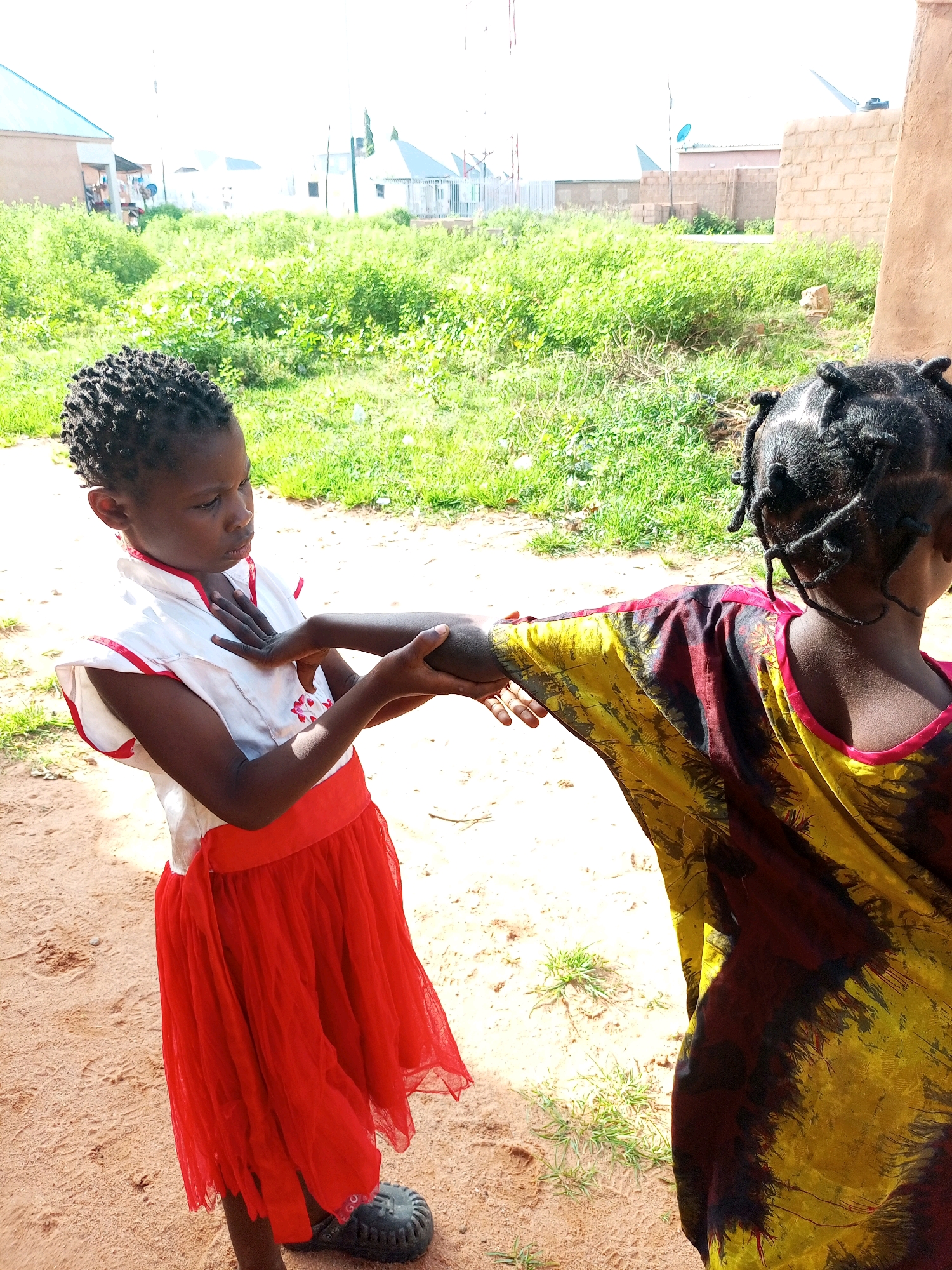 In Tempure, Nigeria, two young girls are engaged in an interaction outdoors. The girl on the left, wearing a white top with red trim and a red skirt, is holding the arm of the girl on the right. The girl on the right is wearing a colorful, tie-dye-like garment in yellow, red, and black. They are standing on a sandy surface with patches of green grass and sparse vegetation. In the background, there are several buildings, some appearing to be under construction, along with more green vegetation under a bright sky. The scene captures a moment of connection between the two children in a semi-urban, outdoor setting.