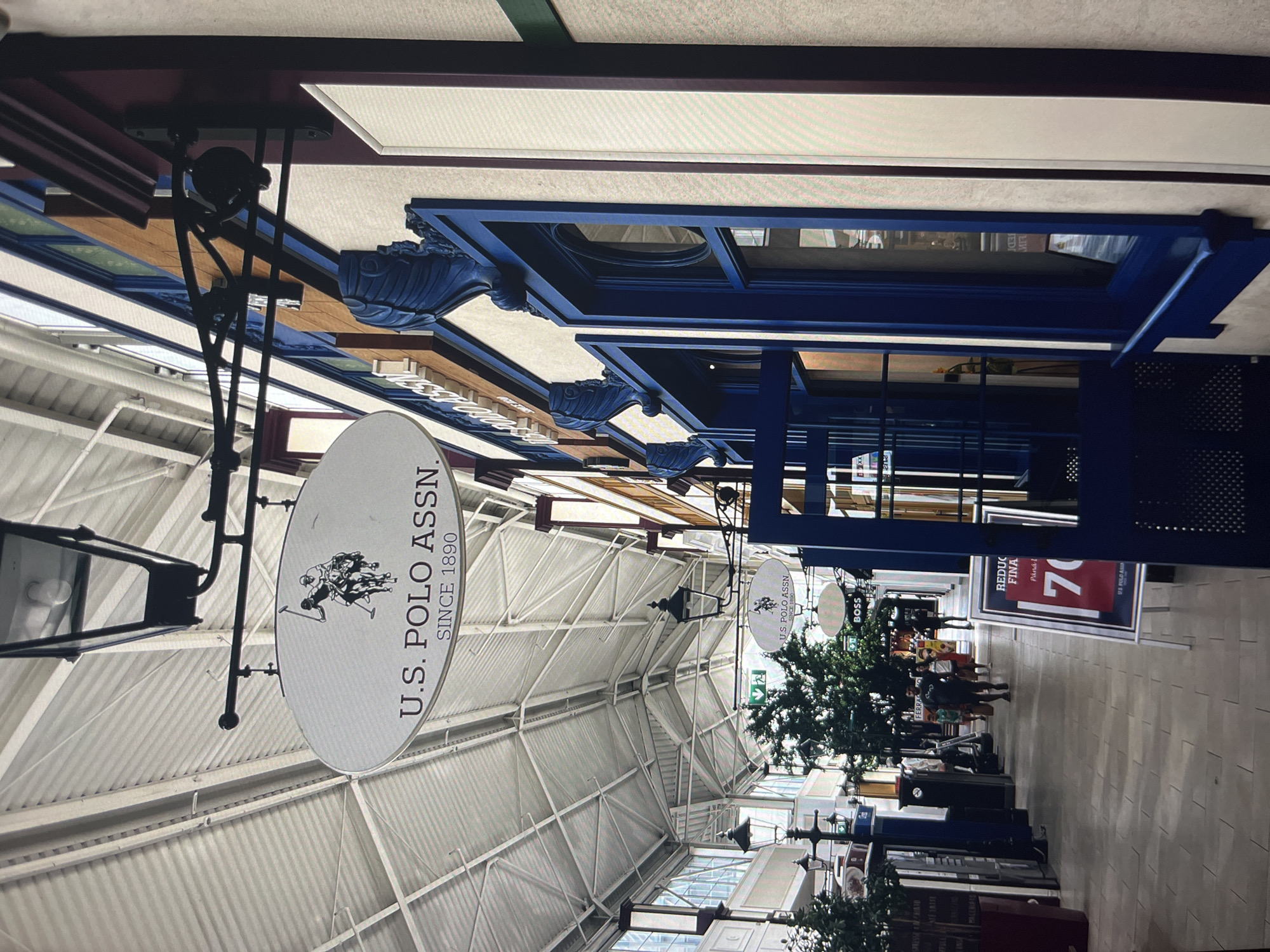 The image shows the interior of a shopping mall in Unknown, Unknown. The mall has a high, white ceiling with metal supports. Hanging signs indicate the presence of stores such as U.S. Polo Assn and Boss. Shoppers are visible in the distance. There is a sign advertising a "REDUCTIONS FINALE" (Final Sale) of 70%. There are also several fake trees spaced evenly within the mall.