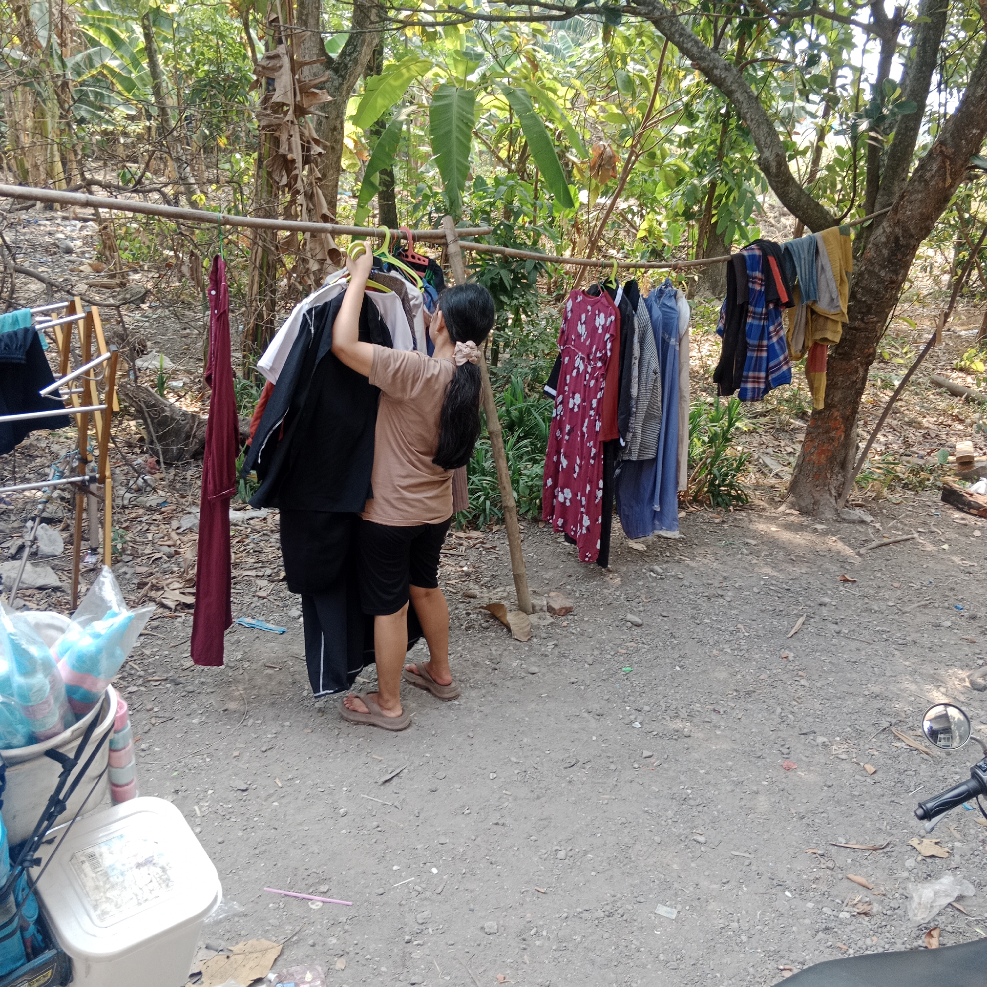 In the village of Tegowanu Kulon, Indonesia, a young woman with dark hair tied in a ponytail is hanging laundry on a clothesline strung between trees and bamboo poles. She wears a light brown t-shirt and black shorts. The clothesline is laden with various garments, including shirts, dresses, and pants, some on hangers. To the left, a metal drying rack holds more clothes. In the foreground, a white plastic bin and a bucket containing items wrapped in plastic are visible on the ground. The scene is set in an outdoor area with a dirt ground, surrounded by vegetation and trees, suggesting a rural or semi-rural environment. The lighting indicates it is daytime, likely sunny.