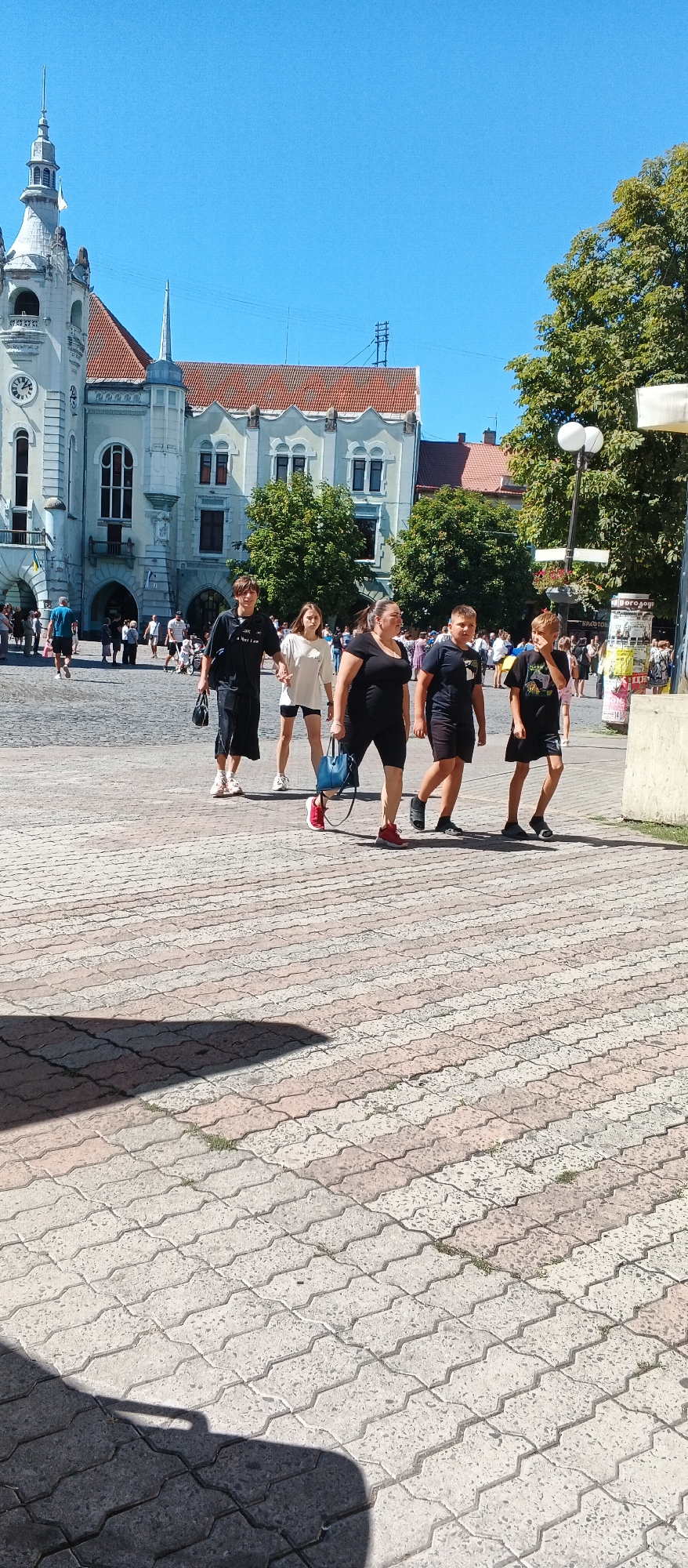 The image depicts a street scene in Mukachevo, Ukraine, with the iconic Palanok Castle in the background. A group of five people, four children and one adult woman, are walking on a cobblestone street. The children are dressed casually in t-shirts and shorts, while the woman is wearing a black t-shirt and black shorts. They appear to be walking together, possibly as a family or group of friends. The street is paved with interlocking bricks, and the buildings surrounding it are old and traditional, with red tiled roofs. The sky is clear and blue, indicating a sunny day. The scene is bustling with activity, with other people visible in the background, suggesting a lively urban environment. The overall impression is one of a typical day in a historic European city.
