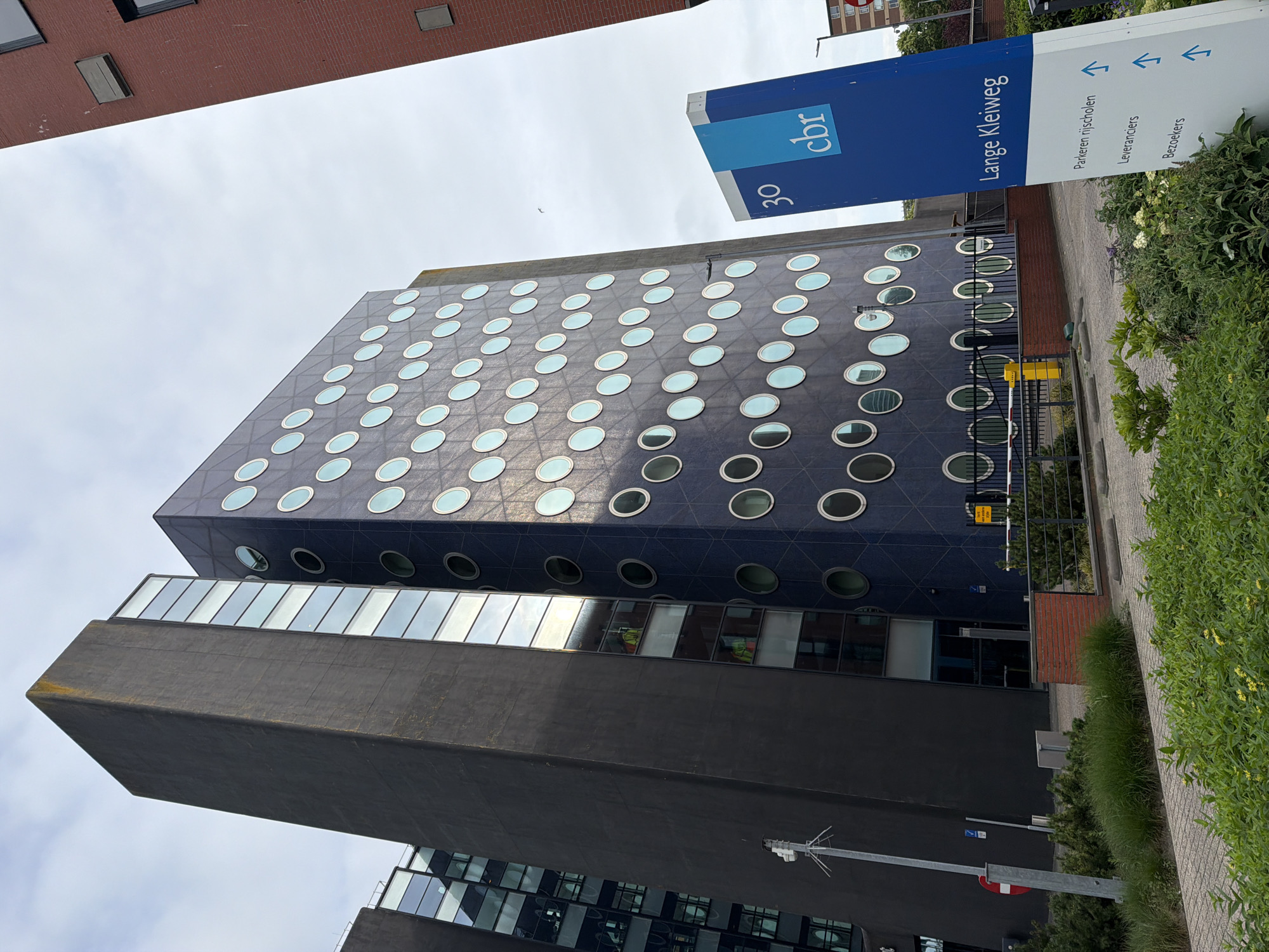 The image shows the CBR building at Lange Kleiweg 30 in Rijswijk, Netherlands. The building is characterized by its dark, angular facade on the left and a blue facade with numerous circular windows on the right. A red brick building is visible to the far right. In the foreground, a blue and white sign indicates "Lange Kleiweg" and lists parking options for "rijrscholen," "leveranciers," and "bezoekers." There are paved areas, a metal fence with a red and white barrier, and lush green vegetation. The sky is overcast.