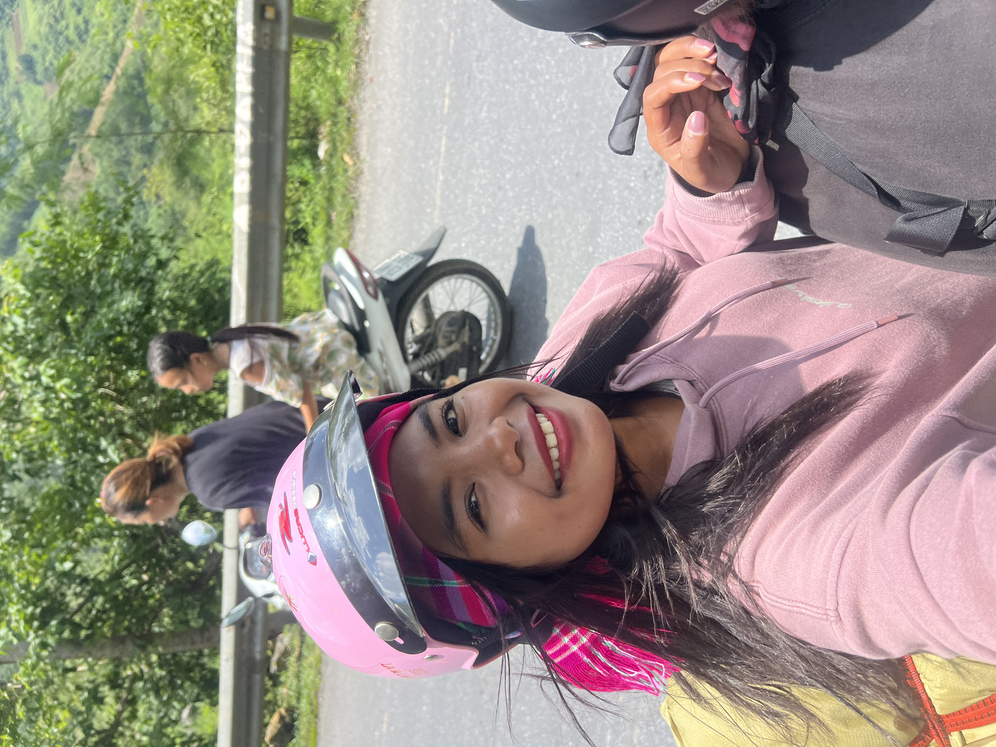 A young woman, wearing a pink helmet and a pink hoodie, smiles at the camera while riding on the back of a motorcycle. She is in Minh Tân Commune, Vietnam. Her hair is long and dark, and she has a pink scarf tied around her head. Another woman and a young girl are on the motorcycle in front of her. The background shows a lush green landscape with trees and mountains. The road is paved, and there is a guardrail on the side.