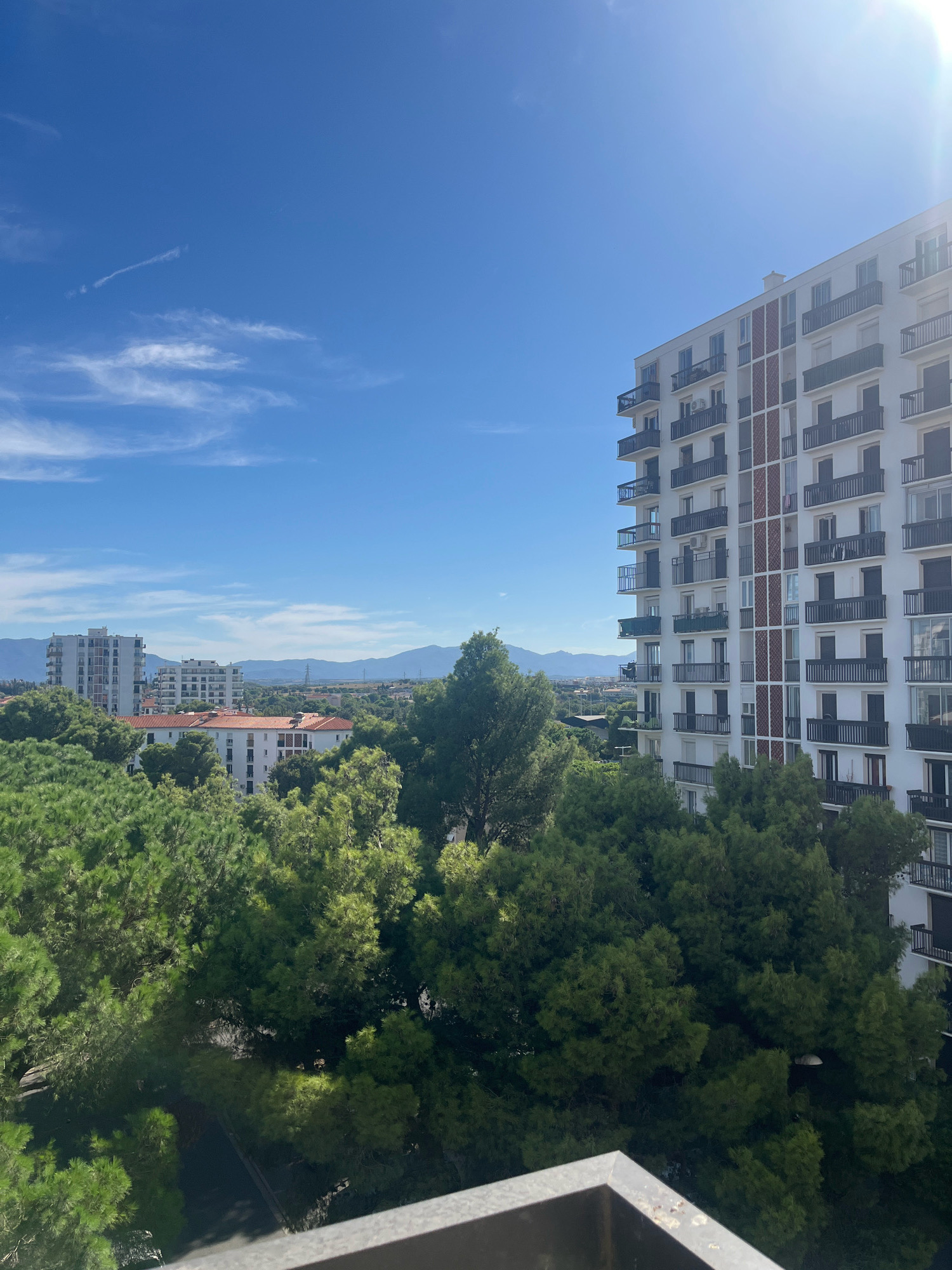 This is a high-angle, wide shot of a city skyline on a clear day with a bright sun. The foreground shows a balcony railing. In the mid-ground, lush green trees fill the frame. Beyond the trees, several multi-story buildings are visible, with one white apartment building in the foreground on the right, featuring numerous dark balconies. In the background, more buildings and mountains can be seen under a bright blue sky with wispy white clouds. The sun is shining from the upper right corner, creating lens flare. There are no people or animals in the image.