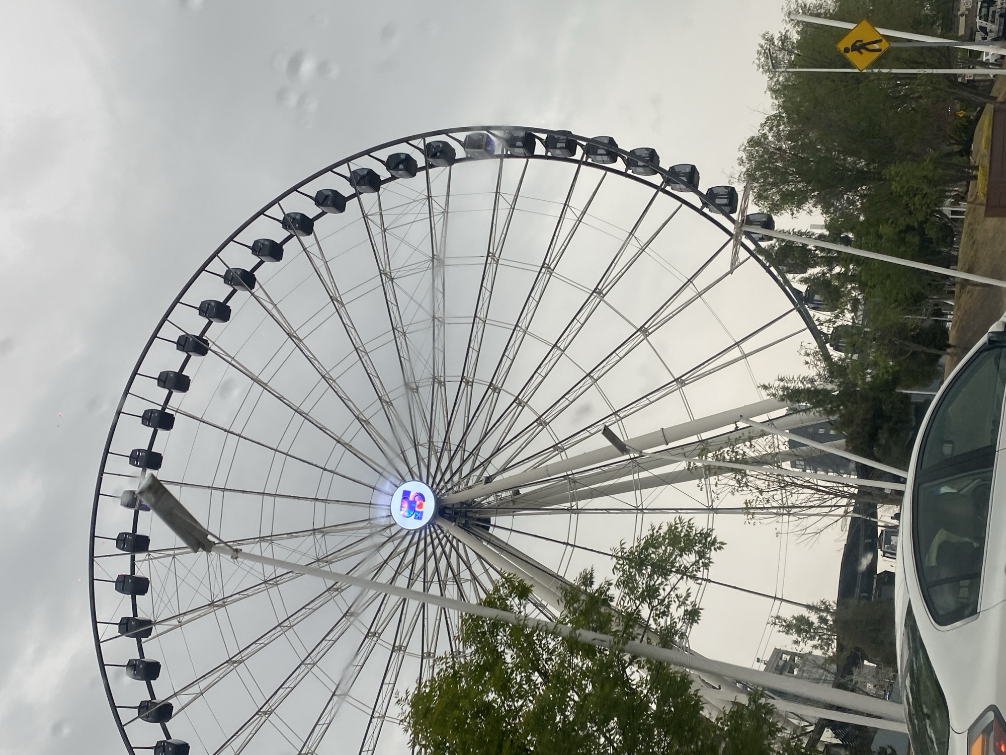 The image shows a large Ferris wheel in Unknown, Unknown. The Ferris wheel has many passenger cars attached around the circumference. The sky is overcast and gray. There are trees and a crosswalk sign visible in the background. A portion of a white car is visible in the lower right corner. The center of the Ferris wheel has a circular logo on it. Raindrops are visible on the windshield of a car.