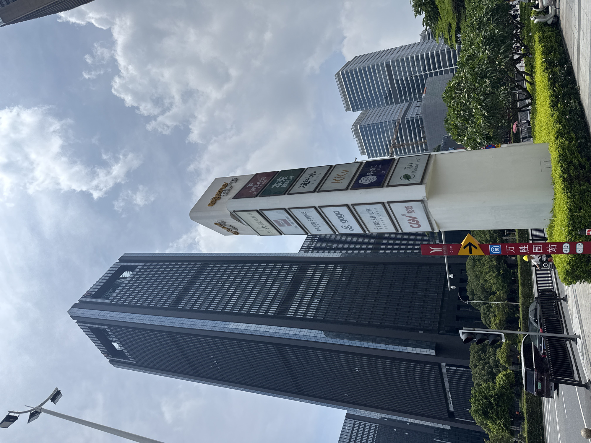 The image displays a modern urban street scene in Haizhu District, China, under a partly cloudy sky. A tall, dark-colored skyscraper with a distinctive top featuring rectangular cutouts dominates the left side of the frame. To its right stands a white, multi-panel commercial directory sign, topped with the "usquare" (悠方) logo, listing various establishments including gaga, T FRESH, KKV, CGV, PAPER STONE, and BENLAI. Further to the right, two additional modern high-rise buildings with layered, curved facades are visible, with a construction crane positioned between them.
In the foreground, a multi-lane road is present with a traffic light showing a red signal. A dark SUV and a black sedan are on the road. A person wearing a yellow hard hat is seen on a white scooter beside a red vertical street sign. This sign features a "Y" logo, "100m," a yellow right-arrow symbol, a bus stop icon, and Chinese characters identifying "万胜围站" (Wanshengwei Station) along with bus route numbers 4, 21, 8, and 28. Lush green landscaping, including hedges and trees, lines the sidewalk and road median. Another individual with a pink umbrella is discernible further down the sidewalk. A white scooter is parked on the paved area near the landscaping on the right. A tall street lamp stands on the far left of the frame.