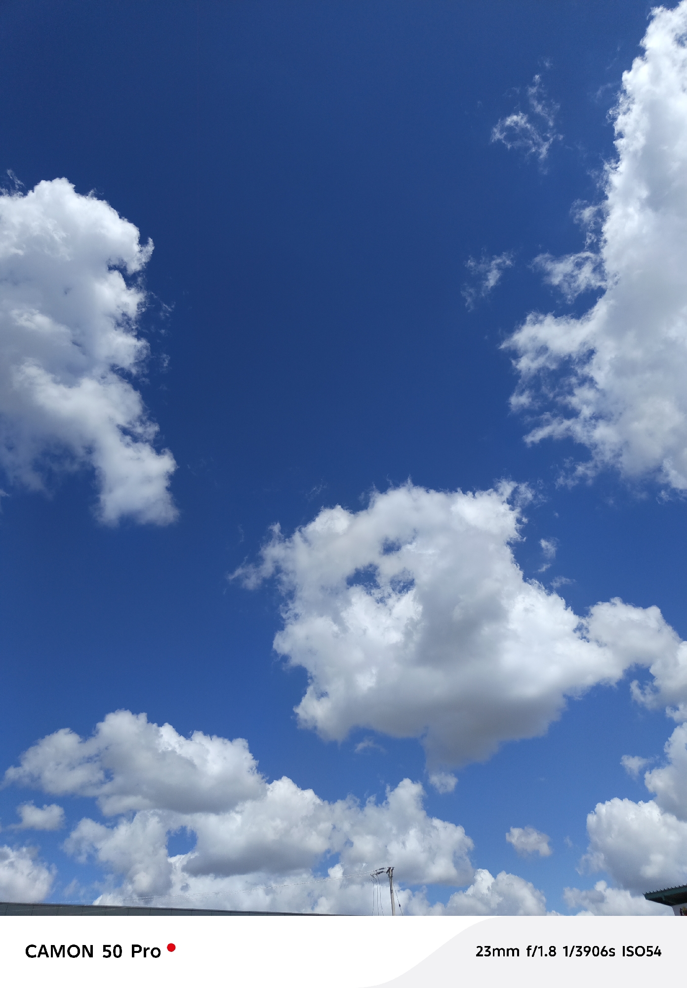 The image shows a bright blue sky with scattered cumulus clouds. In the foreground, a rooftop and a utility pole with wires are visible. The location is likely Dar es Salaam, Tanzania, given the camera model is mentioned as the Camon 50 Pro. No people are visible. The scene is static and depicts a sunny day.
