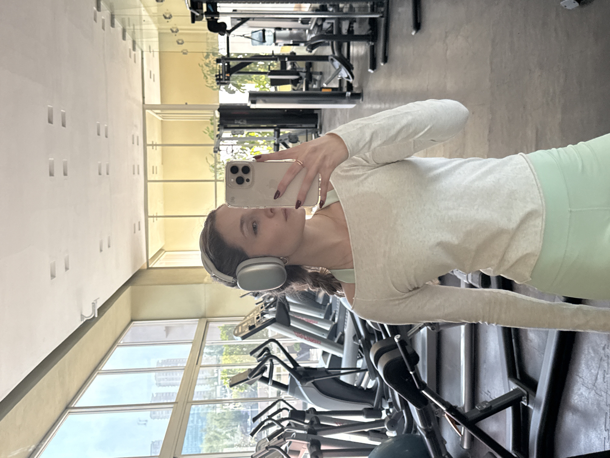 A woman with dark hair, wearing a gray sweatshirt and light green leggings, is taking a selfie in a gym. She is holding a light-colored smartphone in her right hand, partially obscuring her face. She is wearing gray over-ear headphones.
The gym contains various exercise machines, including treadmills and weight machines. Large windows behind the treadmills provide a view of a city skyline, with tall buildings and greenery. Based on the architecture and vegetation visible through the windows, the location is inferred to be in Mexico City, Mexico. The gym's interior is brightly lit.