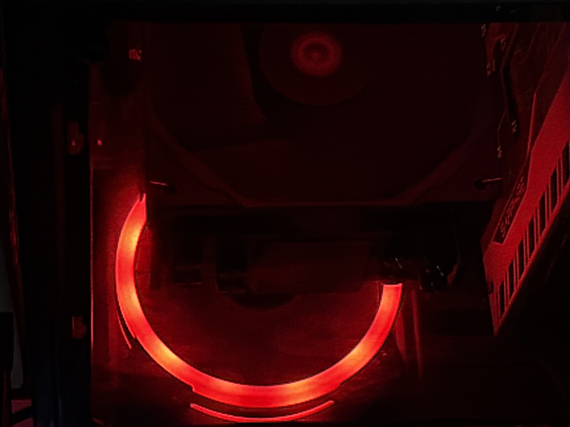 This image depicts the interior of a computer case, located in Moscow, Russia. The scene is dominated by a large circular cooling fan positioned on the left, illuminated by a bright red LED ring around its circumference. The fan blades are discernible but blurred, indicating potential motion. To the right, another dimly lit circular component, possibly a second fan or part of a cooler, exhibits a faint central red glow. Below these, a segment of a circuit board, likely a graphics card or motherboard, is visible, featuring various dark electronic components and heatsink elements, all bathed in the pervasive red light. The overall environment is dark and enclosed, consistent with the inside of a powered-on computer chassis. No individuals, specific actions, or direct interactions are visible within the frame.