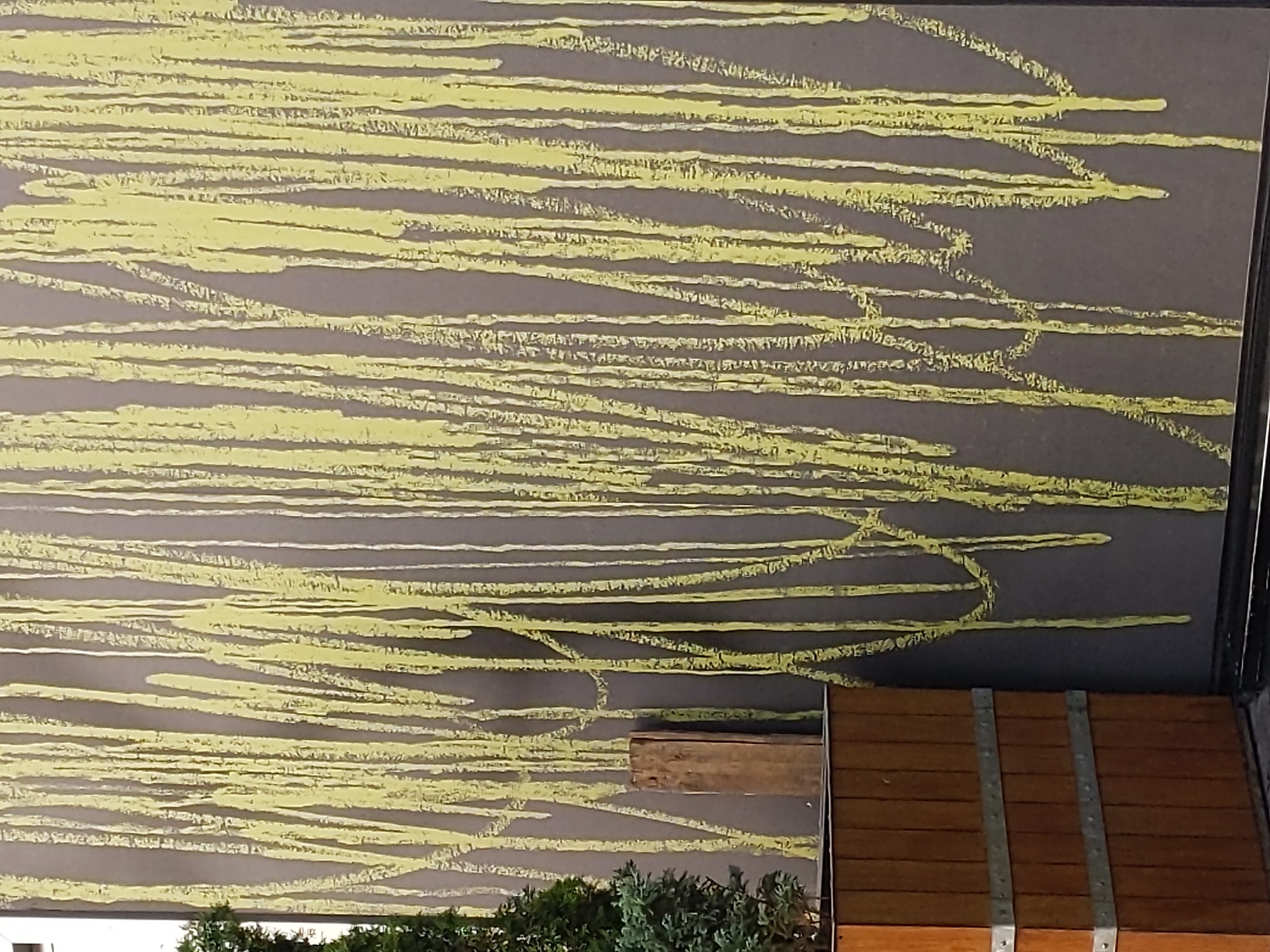 The image shows a section of an exterior wall with a painted mural of yellow vertical lines, some of which are dripping. The mural has a rough texture and appears to be on a dark gray surface. To the left of the mural is a wooden planter box, reinforced with metal bands, containing some greenery. A small wooden post stands next to the planter box, in front of the mural. The scene is likely located in Kyiv, Ukraine, given the context of the request. No people are visible in the image. The overall impression is one of an urban outdoor space with a decorative art element.